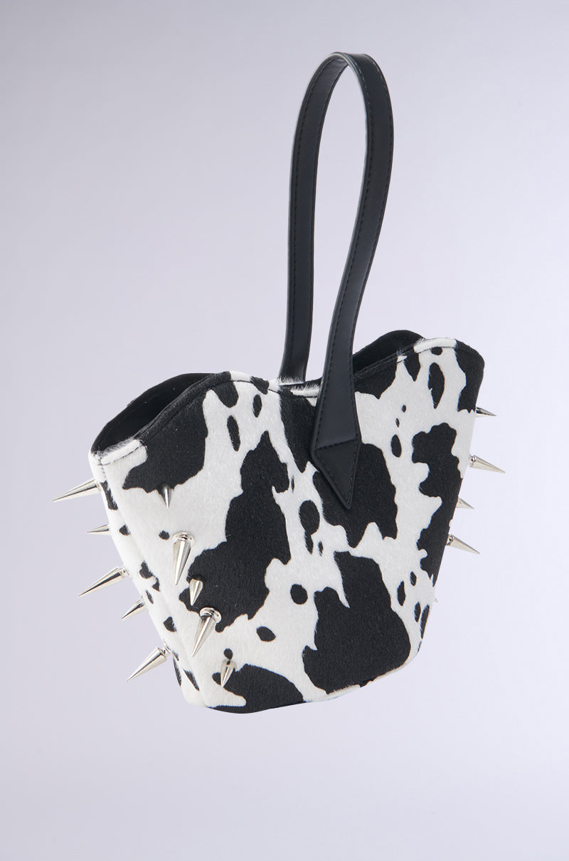 The BUT THIS AIN'T THE FARM MINI PURSE features a cow-print design, black handles, silver spikes, and a chic vegan leather wrist strap—perfect as a statement accessory against any plain light background.