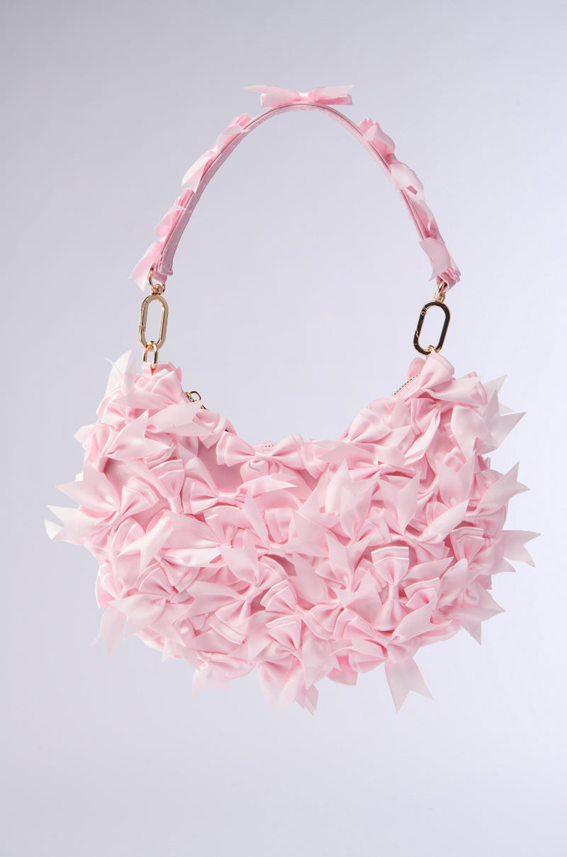 Discover the BOWTIFUL PURSE: a charming baby pink handbag adorned with small bows, featuring a top handle with matching bows and gold clasps. Set against a plain light gray background, this elegant purse adds a playful touch to your style.