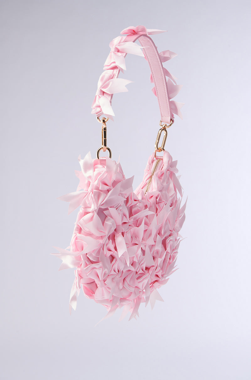 The BOWTIFUL PURSE features petal-like embellishments for a floral look in soft hues and includes a baby pink bow. A top handle with gold-tone hardware contrasts beautifully against its simple gray gradient background.