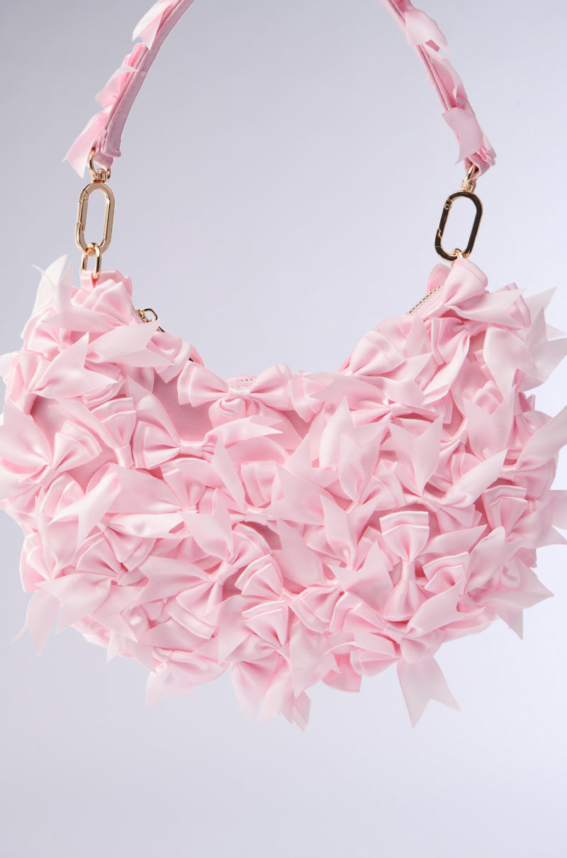 The BOWTIFUL PURSE is a chic handbag featuring a top handle and embellished with baby pink fabric bows connected by gold accents, set against a backdrop that transitions from light gray to white.