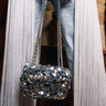 A person in light-wash jeans and white fringe holds the ALL NIGHT LONG SEQUIN PURSE with a chain strap, standing indoors against a blurred brick wall background.