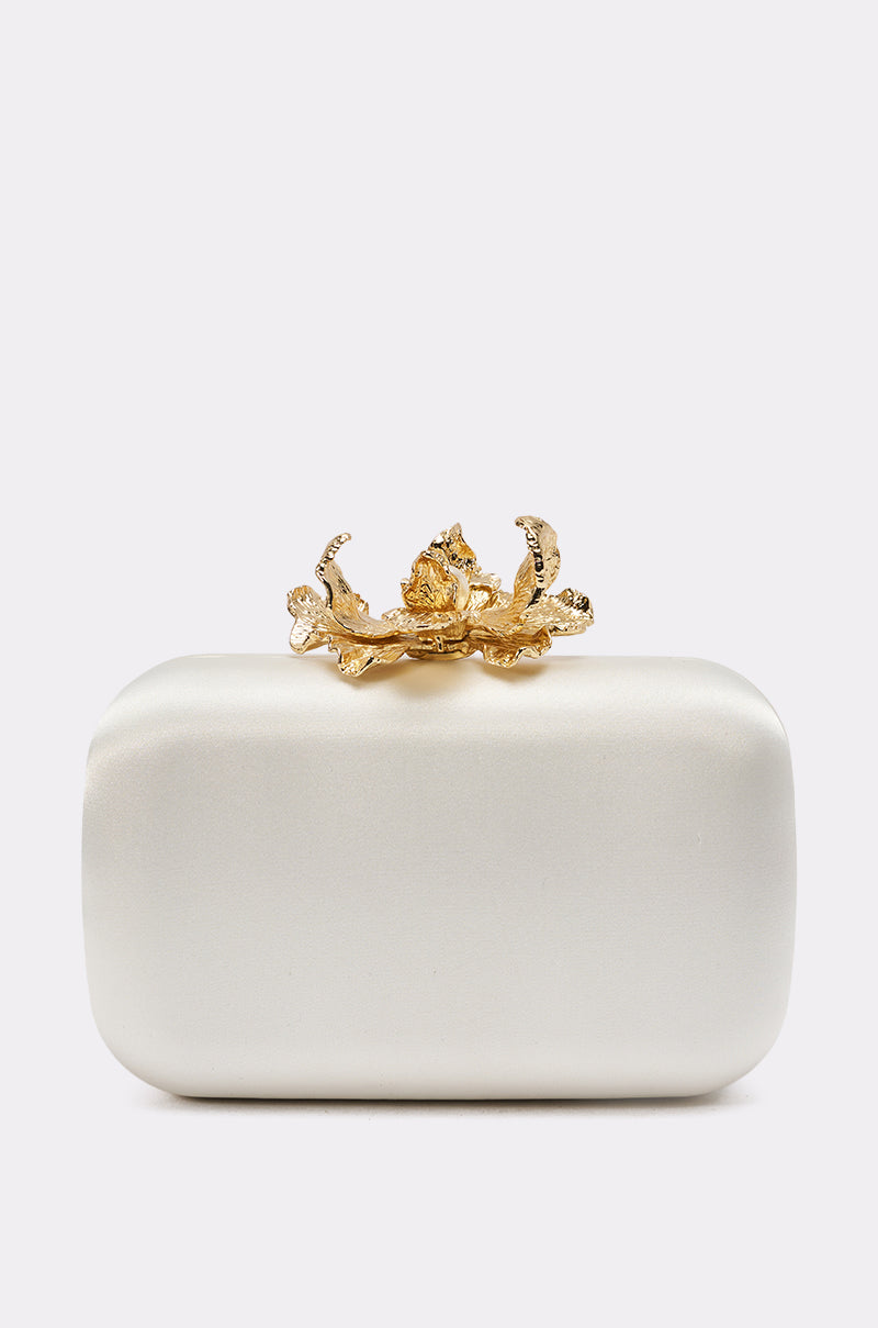 The IN ROME CLUTCH is a rectangular white satin bag featuring a decorative gold floral clasp on top, displayed against a plain light background. 0