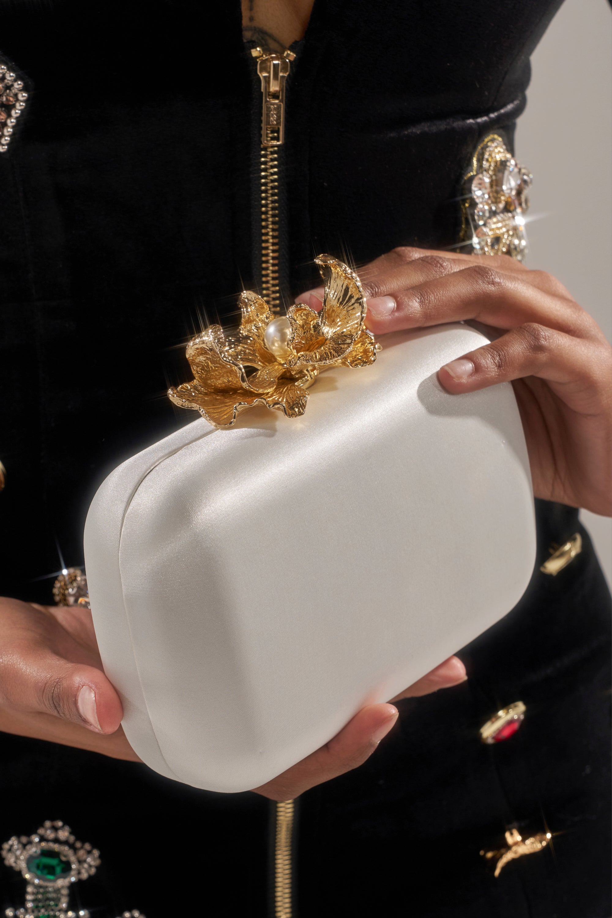 A person in a black jeweled outfit holds the IN ROME CLUTCH, a structured white satin bag featuring an ornate gold and pearl flower-and-bird clasp with gold-tone hardware.