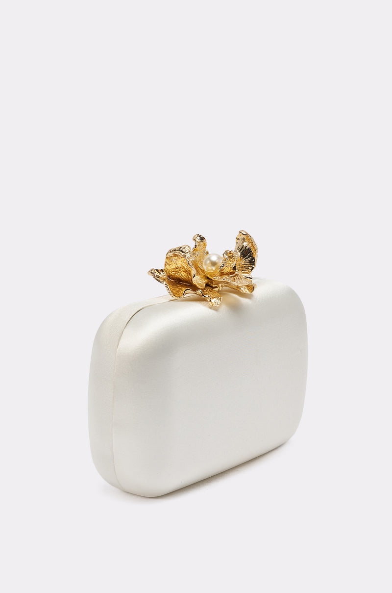 The IN ROME CLUTCH is a small, rectangular white bag with rounded edges and a gold floral clasp, adding structured elegance to any outfit. Set against a plain light background. 3