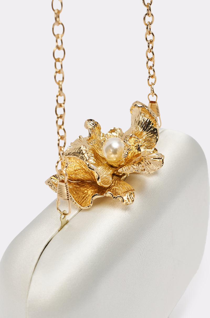 The IN ROME CLUTCH is a structured white handbag featuring a removable gold chain strap and an elegant gold flower with a pearl center on top.