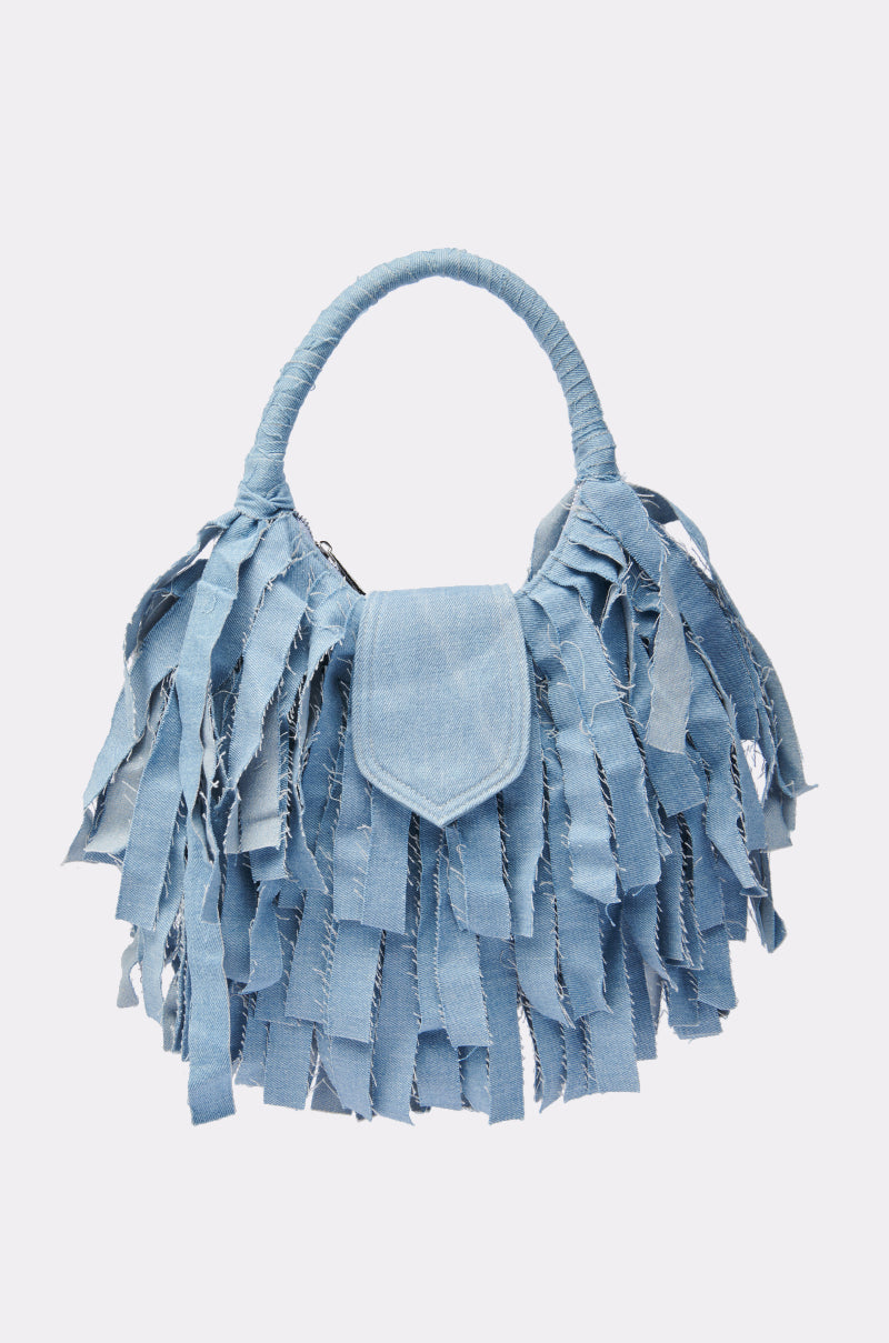 The SUZIE DENIM FRINGE PURSE is a light blue denim handbag with a rounded handle and multiple fringed strips, creating a textured and playful look.