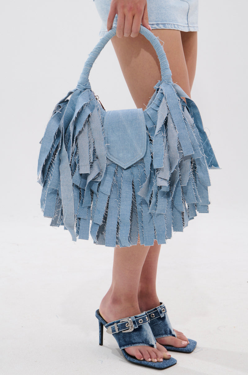 A person stands on a white floor wearing a light denim mini skirt and denim high-heeled sandals, holding the SUZIE DENIM FRINGE PURSE with a short handle for the perfect street-style look.