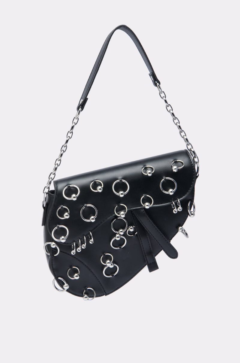 The PIERCE PLEASE PURSE is a black faux leather bag featuring a short chain and leather strap, silver-tone hoop piercings, grommets, and an asymmetrical flap—an edgy-chic accessory perfect for modern style.