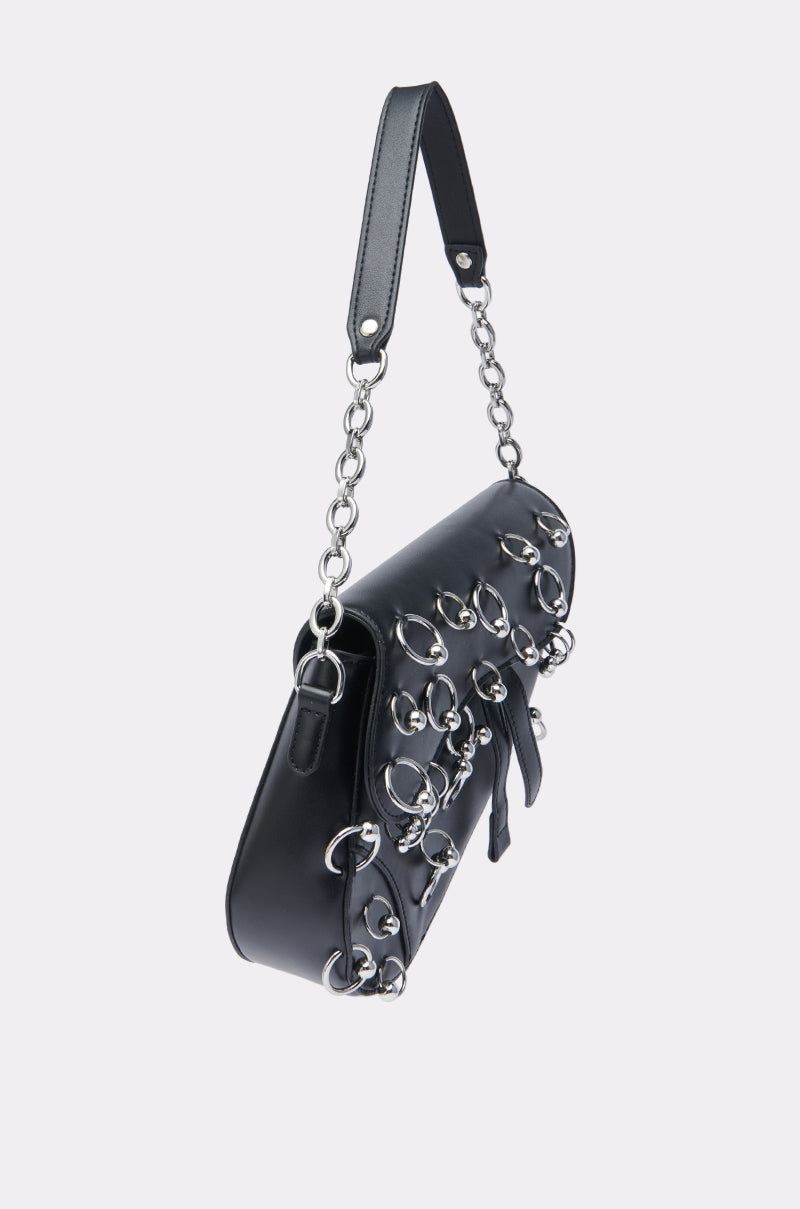 The PIERCE PLEASE PURSE is a black faux leather bag with a short strap and metal chain links, featuring edgy silver-tone hoop piercings and loops on the front. Shown against a plain, light background.