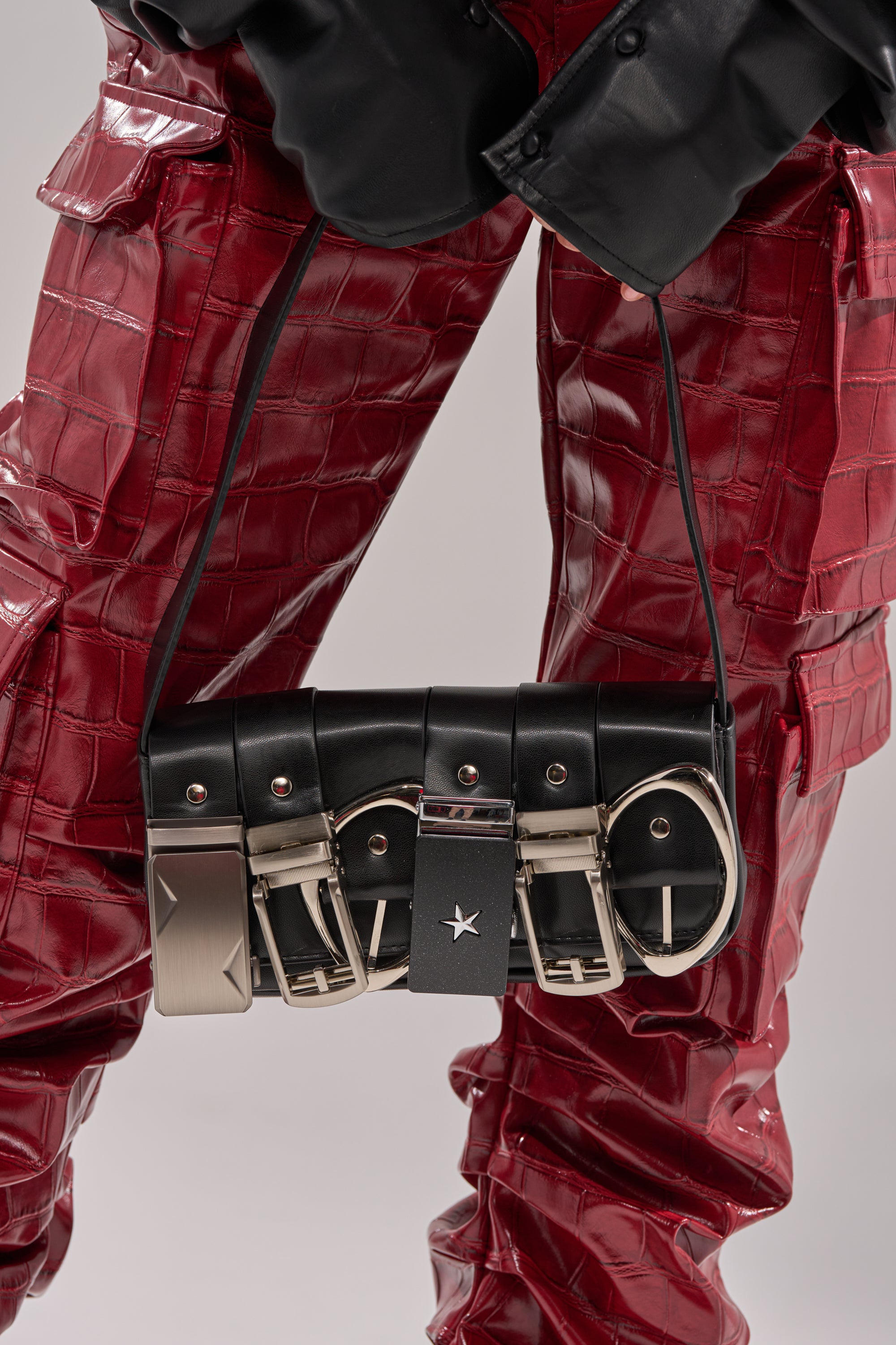 A person in shiny red crocodile-print pants and black gloves holds the BUCKLE UP PURSE, a unique vegan leather bag featuring silver buckle accents and a star charm.
