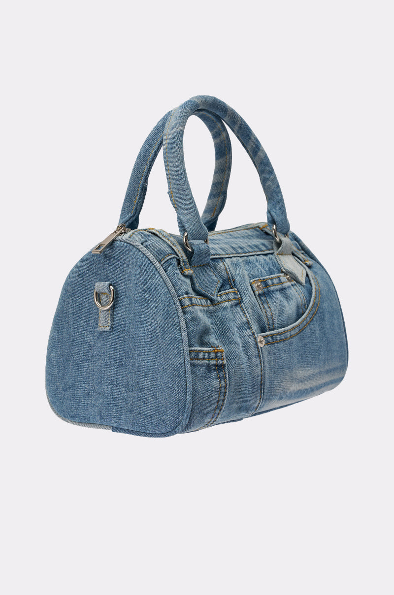 The NO BLUES DENIM PURSE is a mini duffel-style bag crafted from blue denim with jeans-inspired pockets, seams, and metal rivets. It features two short handles, a rounded shape, and a removable strap for versatile carrying. 2