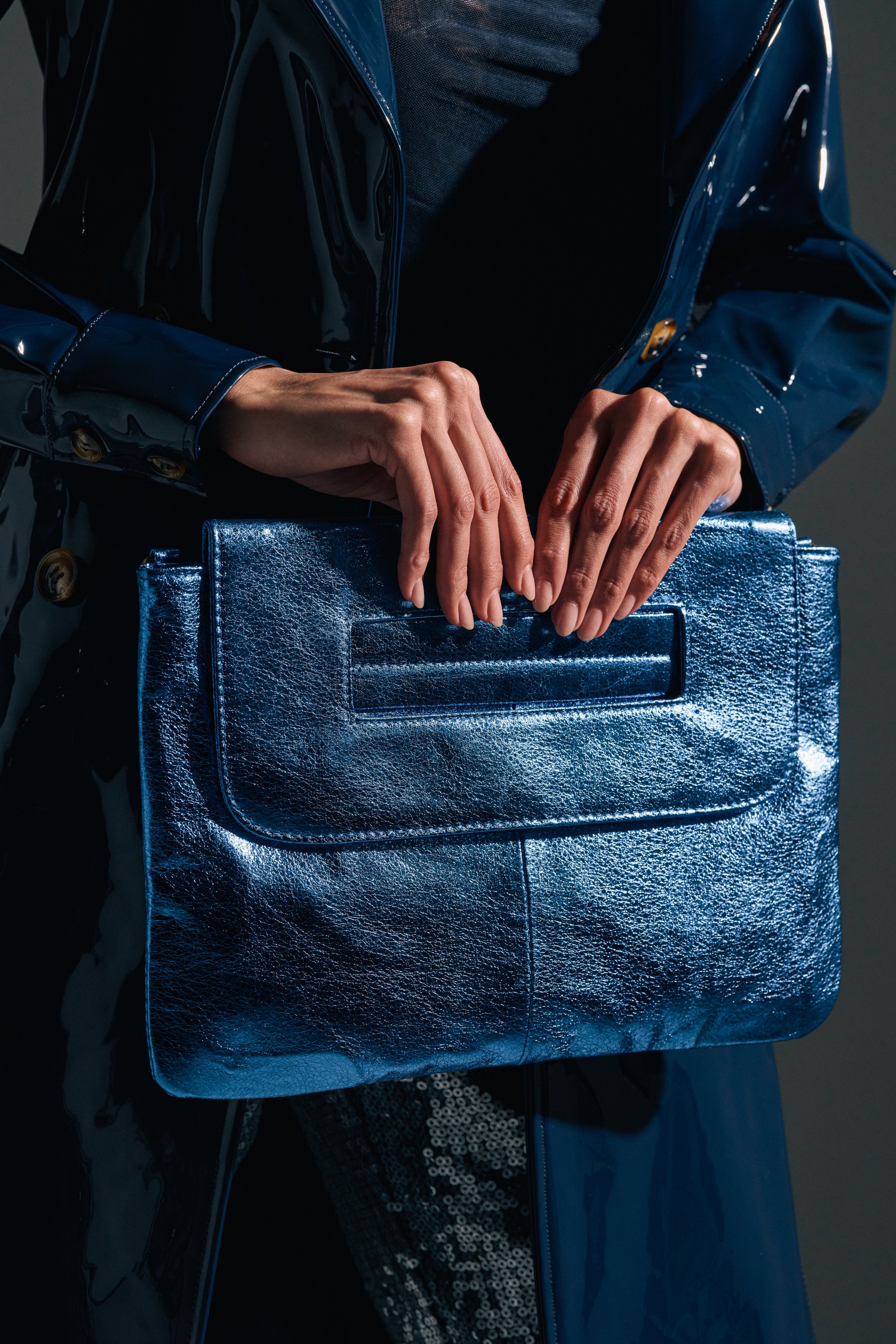 A person in a shiny dark blue trench coat holds the COSMIC COBALT OVERSIZED CLUTCH, its metallic blue finish gleaming in their hands against a dark background.