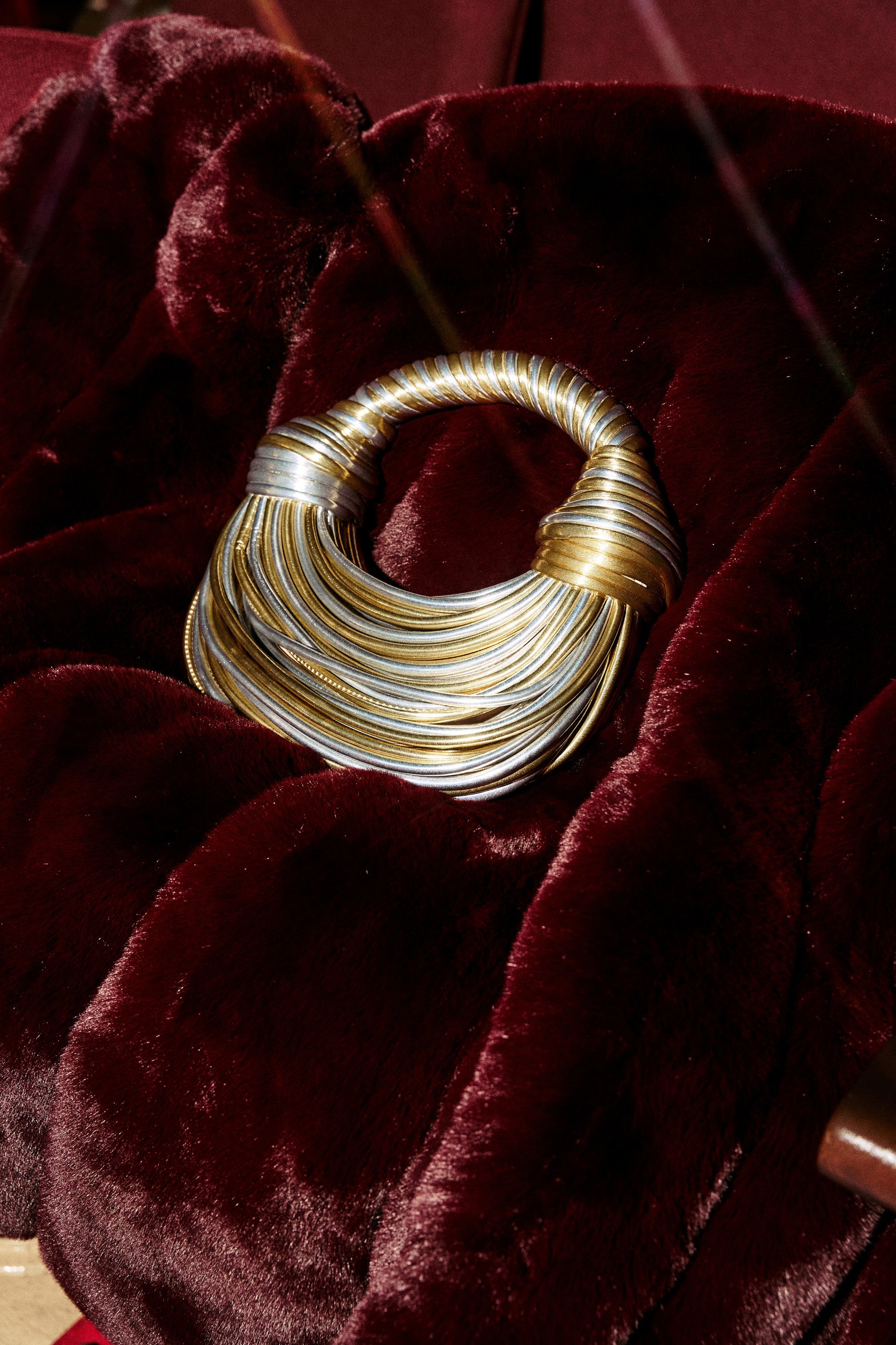 A chunky, metallic gold and silver twisted statement necklace rests on deep burgundy faux fur, styled beside the chic FIND ME OUTSIDE PURSE. 2