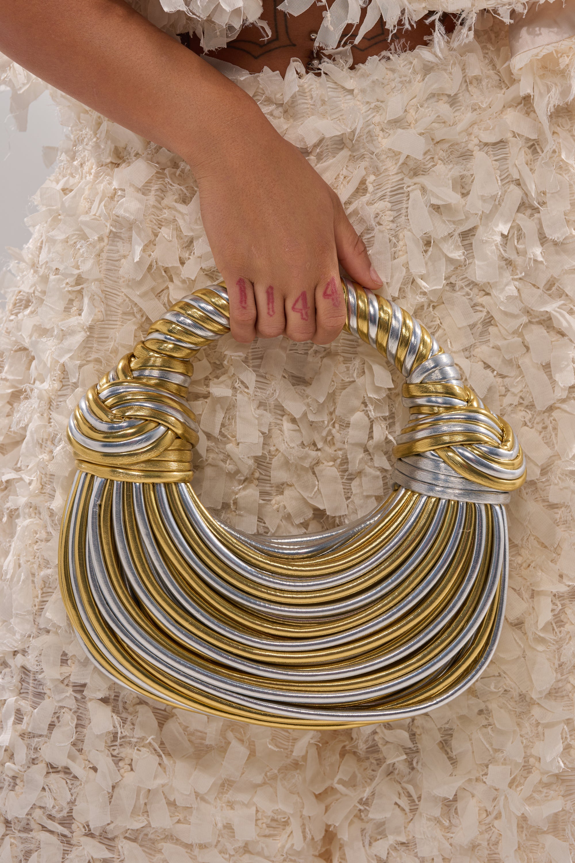 A person wearing a textured cream skirt holds the FIND ME OUTSIDE PURSE, a metallic faux leather bag with gold and silver stripes, a twisted handle, and a unique crescent silhouette. 4