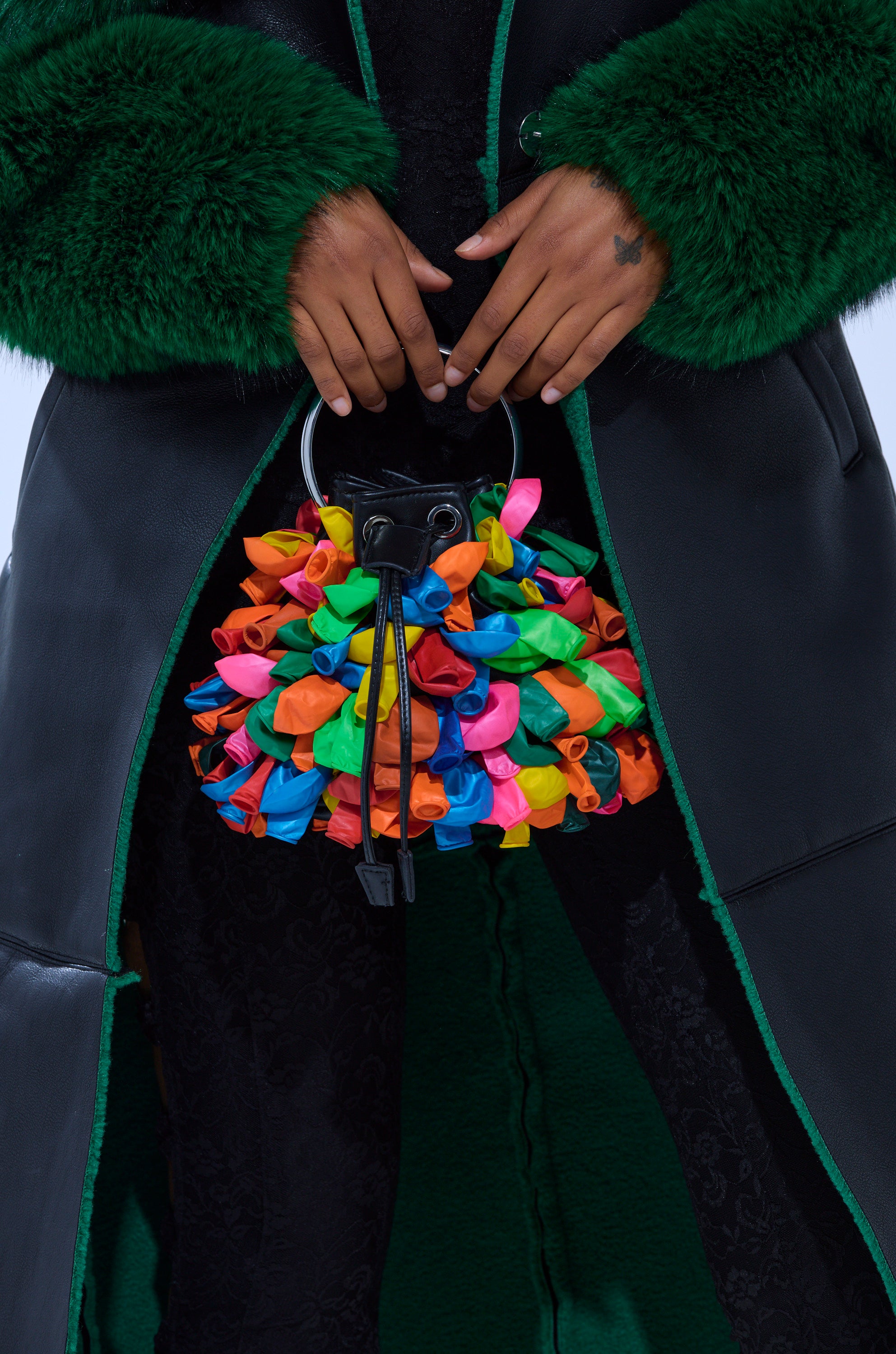 A person in a dark green fur and leather coat holds the BLOW ME BUCKET BAG, a faux leather handbag featuring colorful twisted balloon details. The simple background highlights the playful bag and the luxurious clothing textures.