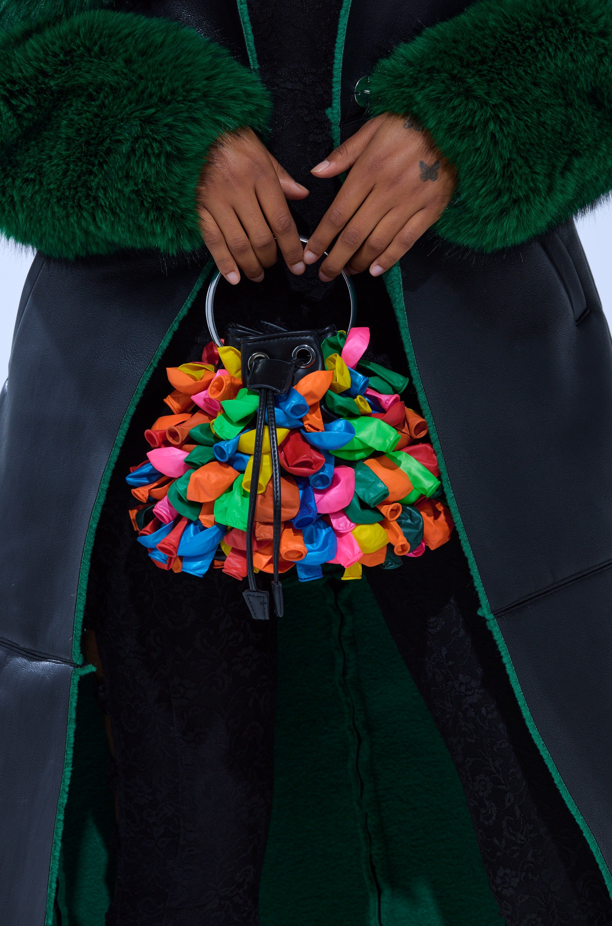 A person in a dark green fur and leather coat holds the BLOW ME BUCKET BAG, a faux leather handbag featuring colorful twisted balloon details. The simple background highlights the playful bag and the luxurious clothing textures. 1