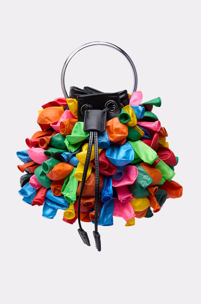 The BLOW ME BUCKET BAG features a round shape, black faux leather accents, a metal handle, and vibrant balloon details in red, green, blue, yellow, orange, and pink for a playful textured look.