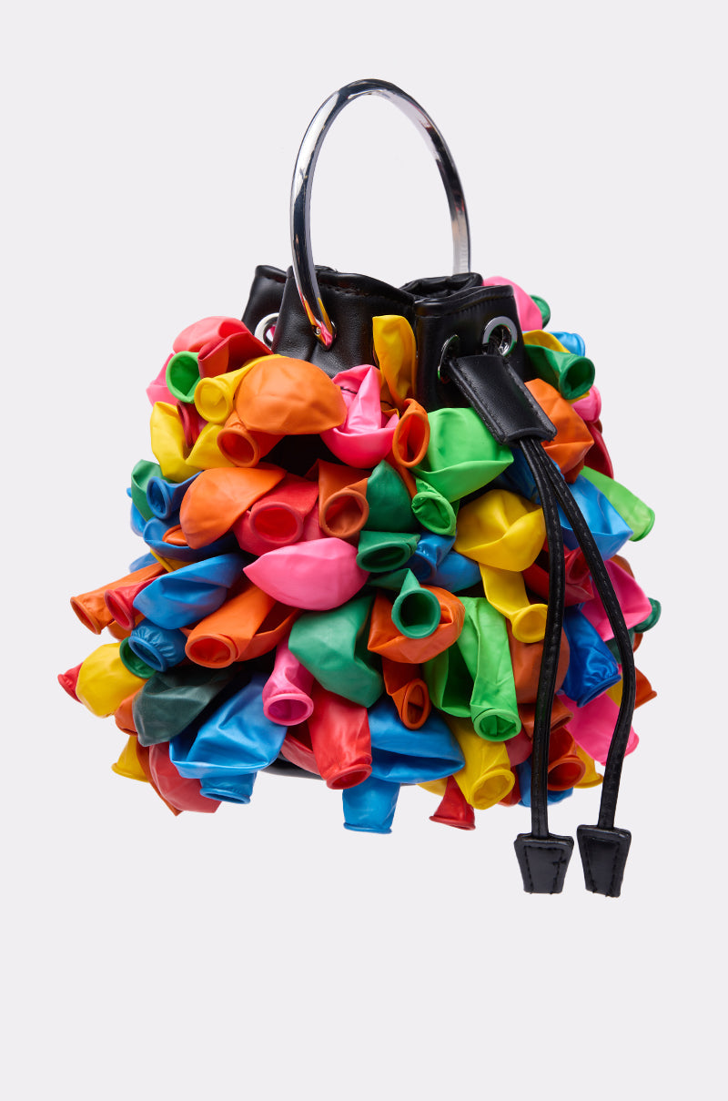 The BLOW ME BUCKET BAG is a black faux leather bucket bag with colorful balloon details, silver handles, and a black drawstring closure, shown on a plain white background. 2