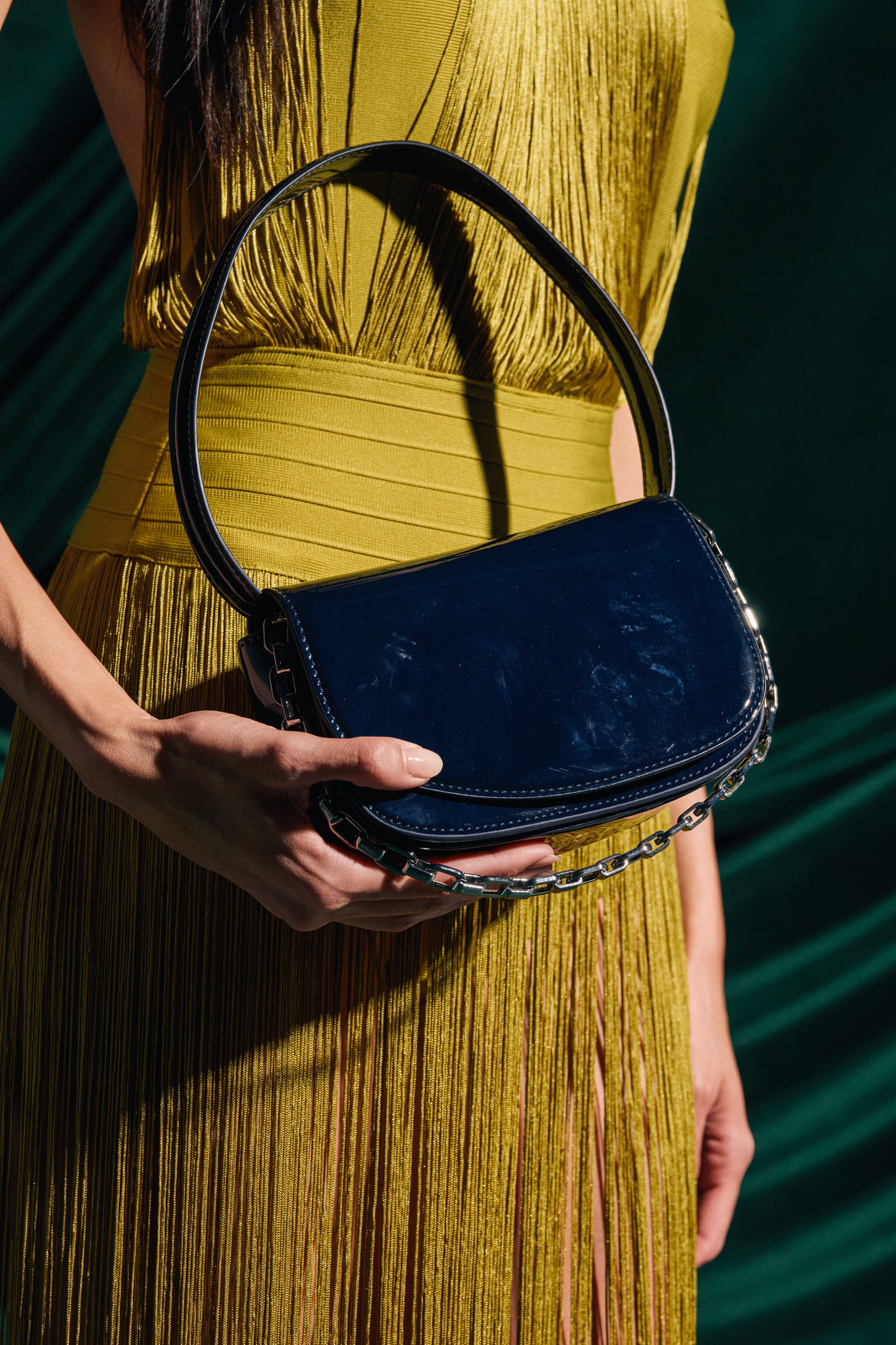 A person in a yellow fringed dress holds the WITH IT PATENT PURSE, a shiny black mini-bag with a curved handle and chain detail, standing against a dark green background. 0