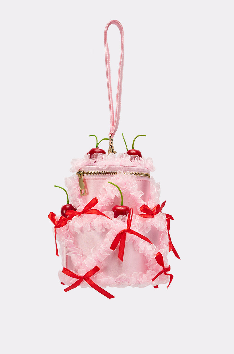 The MARIE CHERRY PURSE is a satin handbag shaped like a pink tiered cake, featuring red bows, ruffled trim, faux cherries, a pink strap, and a gold zipper.