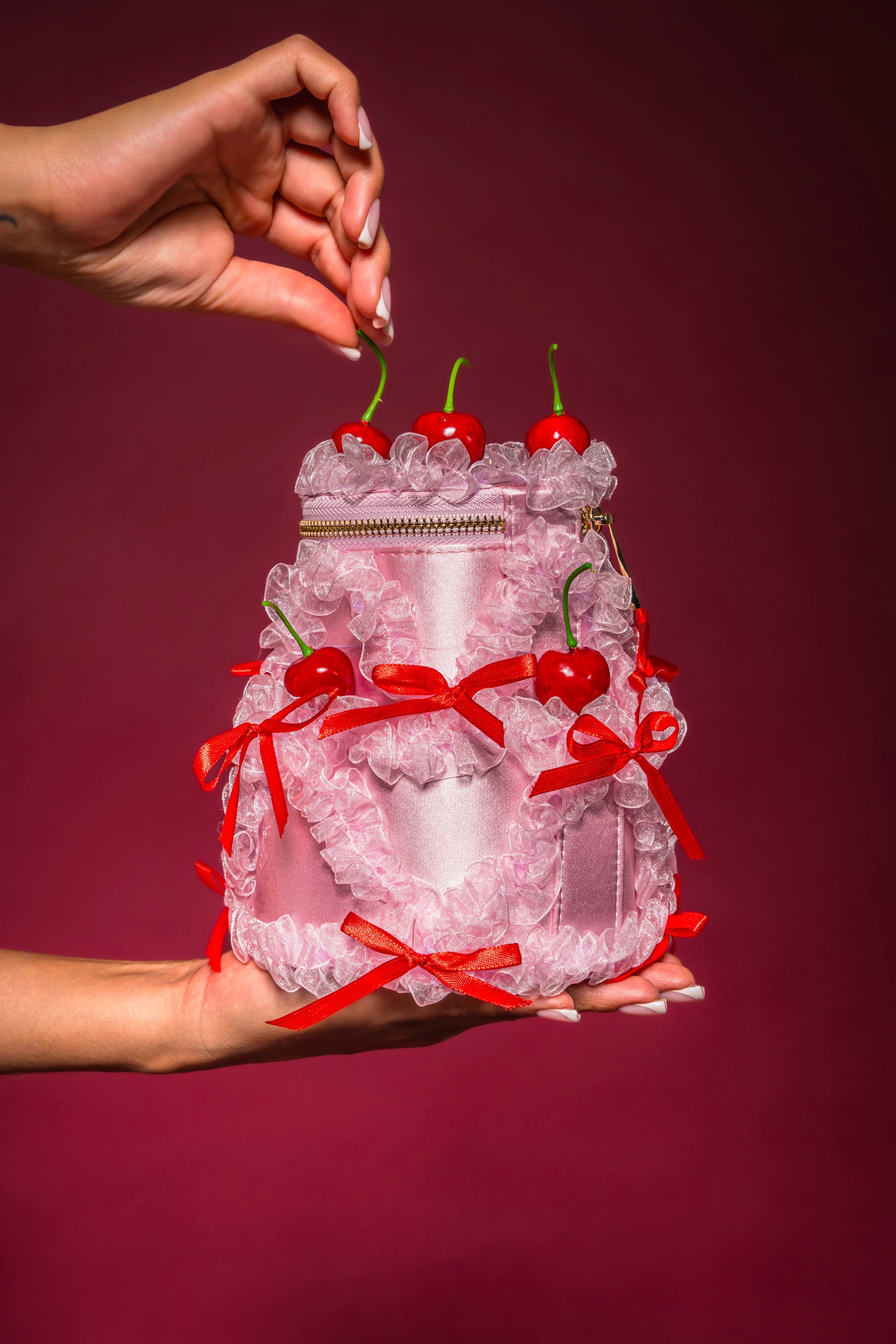 The MARIE CHERRY PURSE, a pink satin handbag shaped like a cake with ruffles, red bows, and faux cherries on top, is held in one hand as another hand reaches for a cherry against a deep burgundy background.