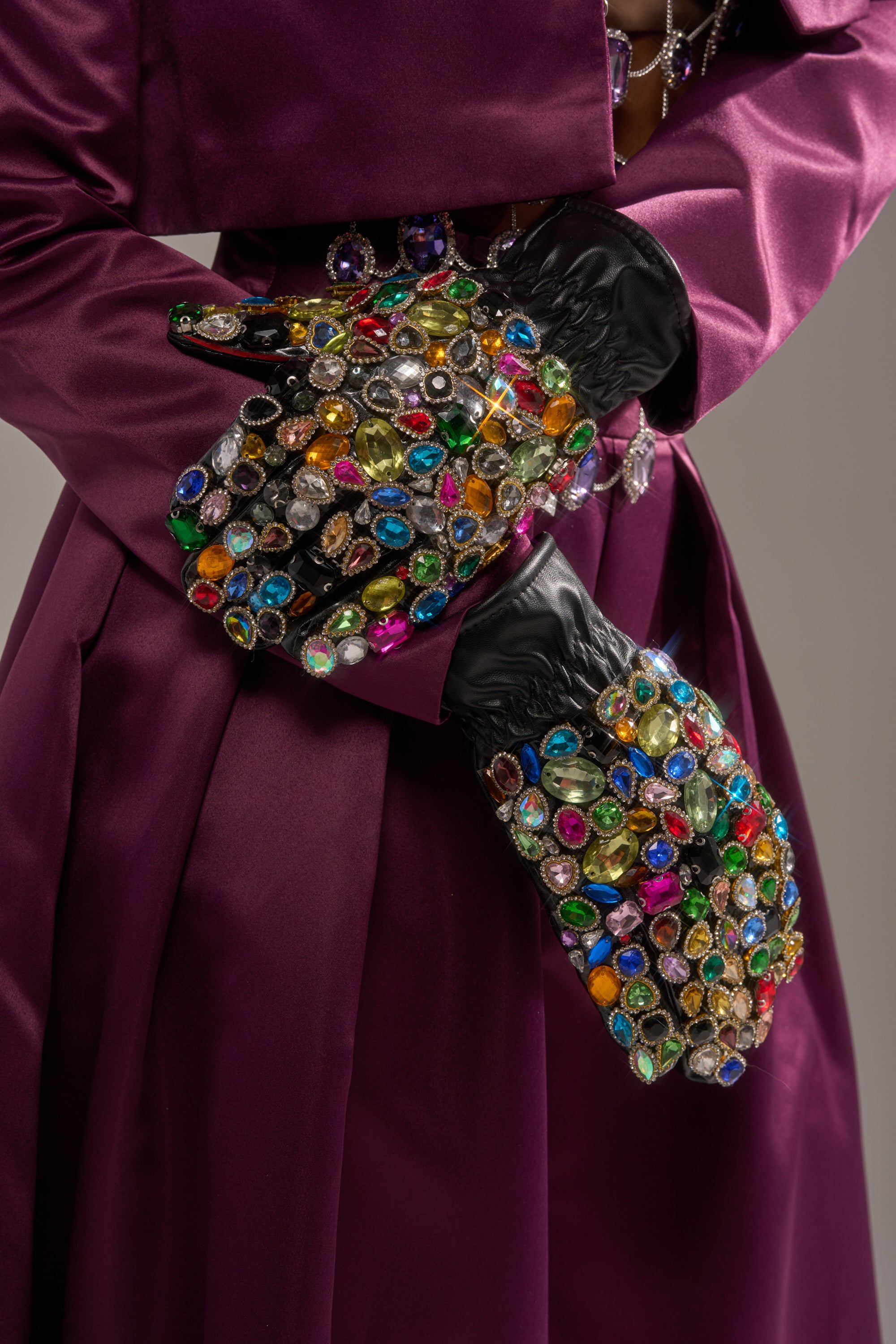 A person in a deep purple satin coat crosses their arms, showing off LOVE YOU BLING GLOVES adorned with large, colorful gem-like stones. The sparkling, ornate gloves stand out against the rich fabric.