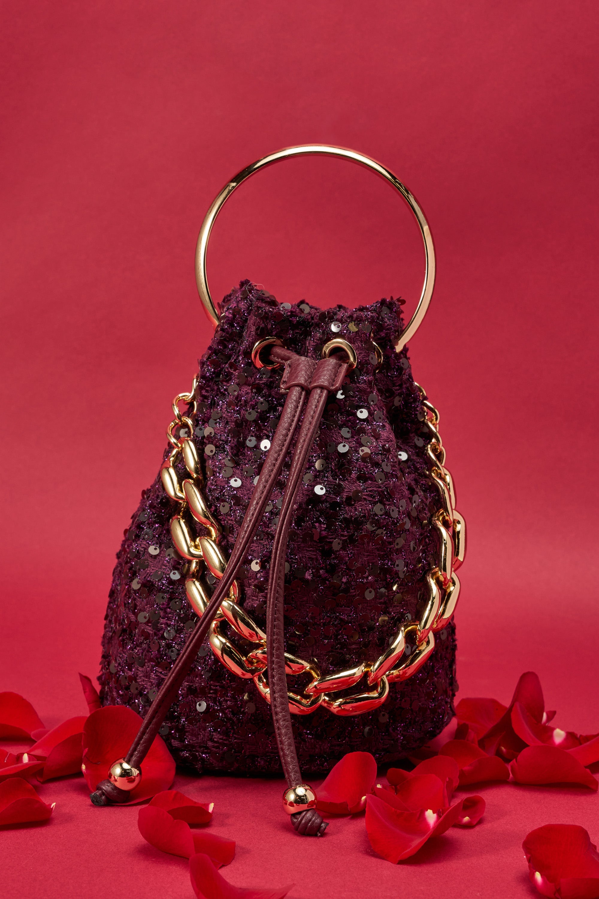 The KARY TWEED BUCKET BAG, a small deep purple mini bucket bag with gold chain accents and a gold-tone handle, is displayed on a red surface with scattered rose petals against a red background. 0