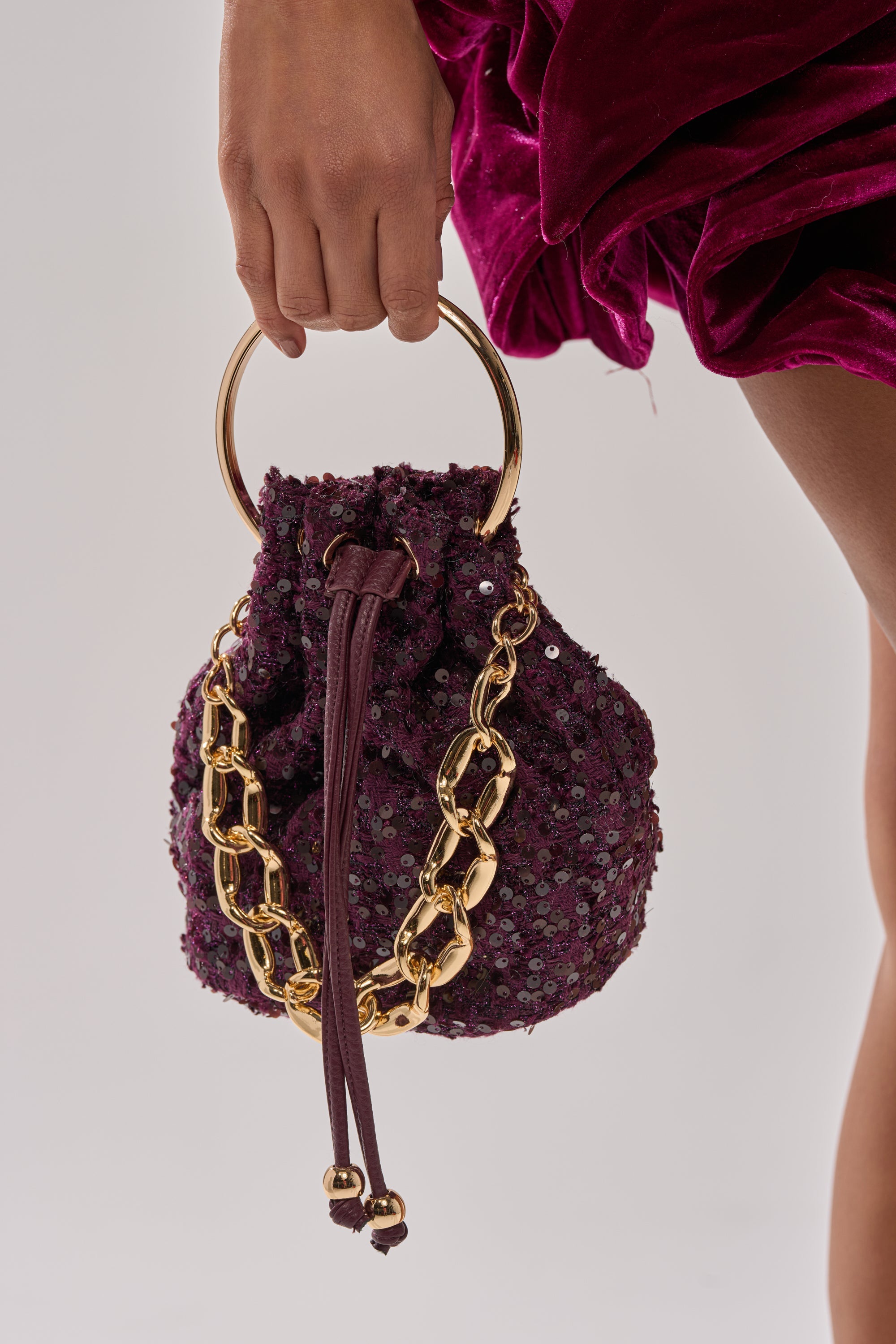 A person in a dark magenta velvet dress holds the KARY TWEED BUCKET BAG, featuring textured purple fabric, a gold-tone top handle, and a chunky gold chain accent. The background is plain and light-colored. 2