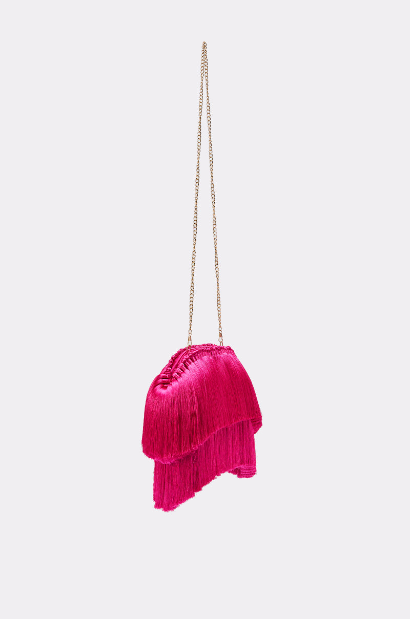 The RAZZLE DAZZLE TASSEL CLUTCH is a small, bright pink fringe clutch with layered tassels and a slim gold chain strap, standing out against a plain light gray background. 3