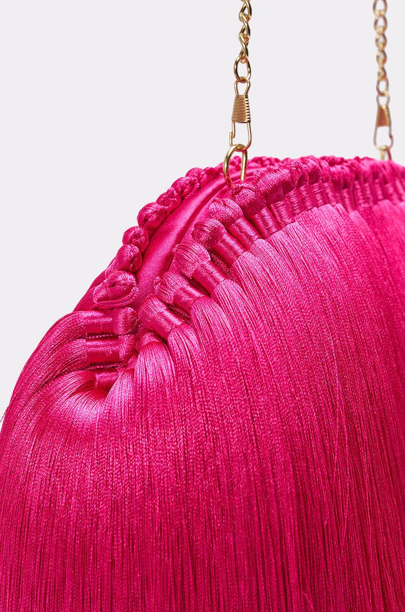 Close-up of the RAZZLE DAZZLE TASSEL CLUTCH, a vibrant pink clutch with silky fringe details and a gold chain strap—an eye-catching statement piece set against a simple, light background. 4