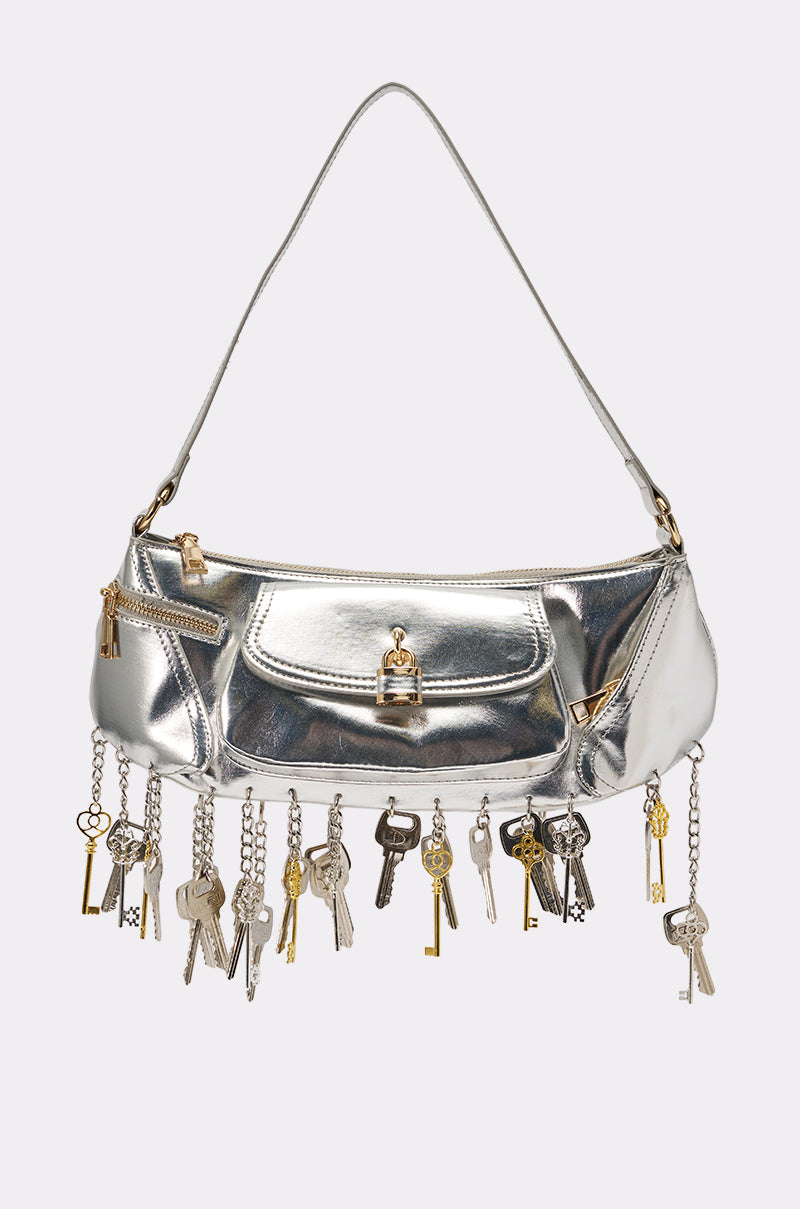 The THATS THE KEY PURSE is a metallic silver shoulder bag with a short strap, front pocket, and gold and silver key charms, safety pins, and keychains hanging as fringe along the bottom edge for an edgy glam look. 1