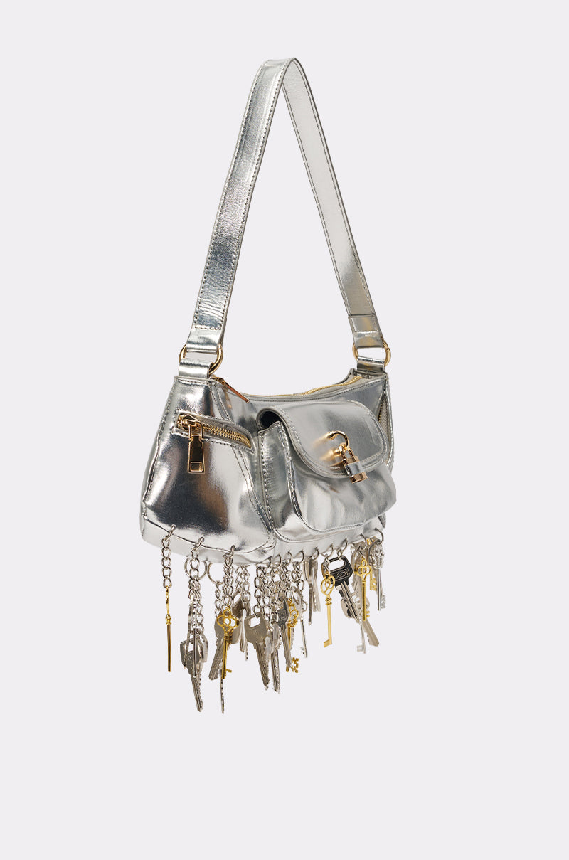 The THATS THE KEY PURSE is a metallic silver shoulder bag with a short strap, gold hardware, and multiple lock and key charms dangling from chains along the bottom edge. 2