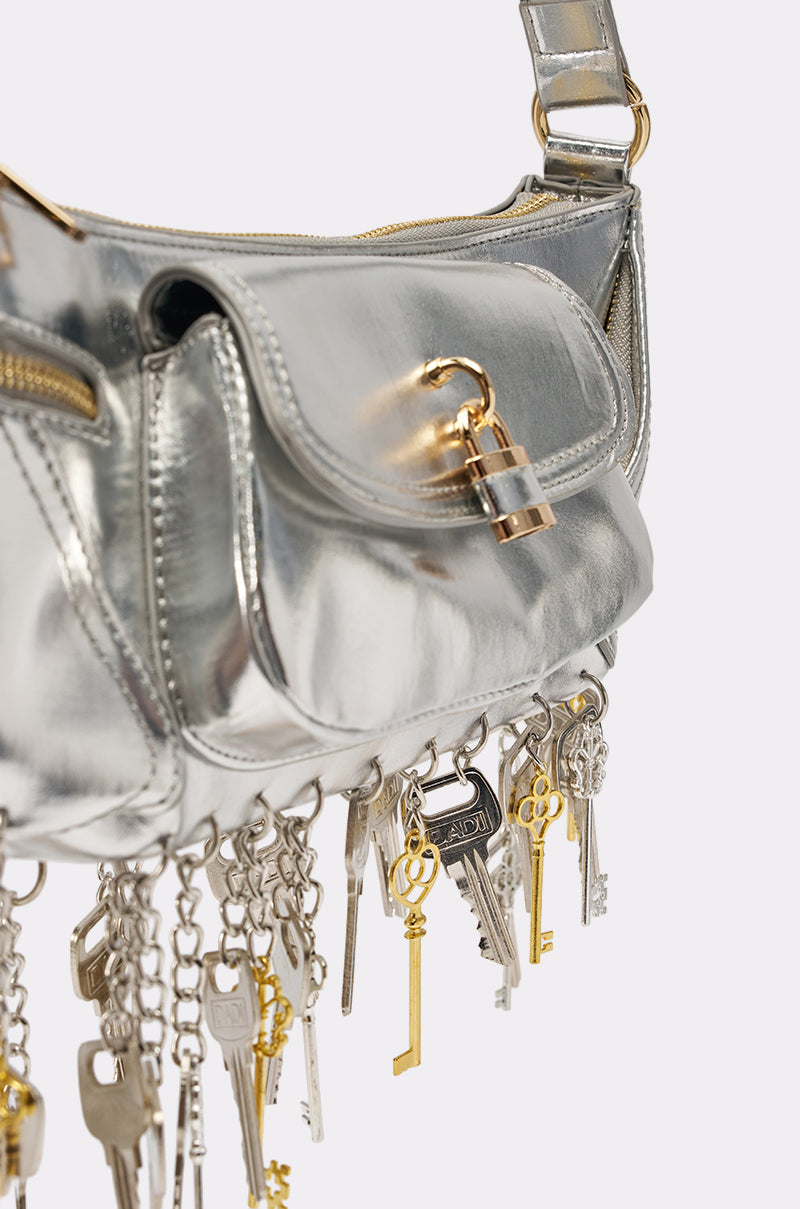 The THATS THE KEY PURSE is a chic metallic silver shoulder bag with a front flap, small gold padlock closure, and a stylish fringe of gold and silver key charms dangling from chains at the bottom. 3