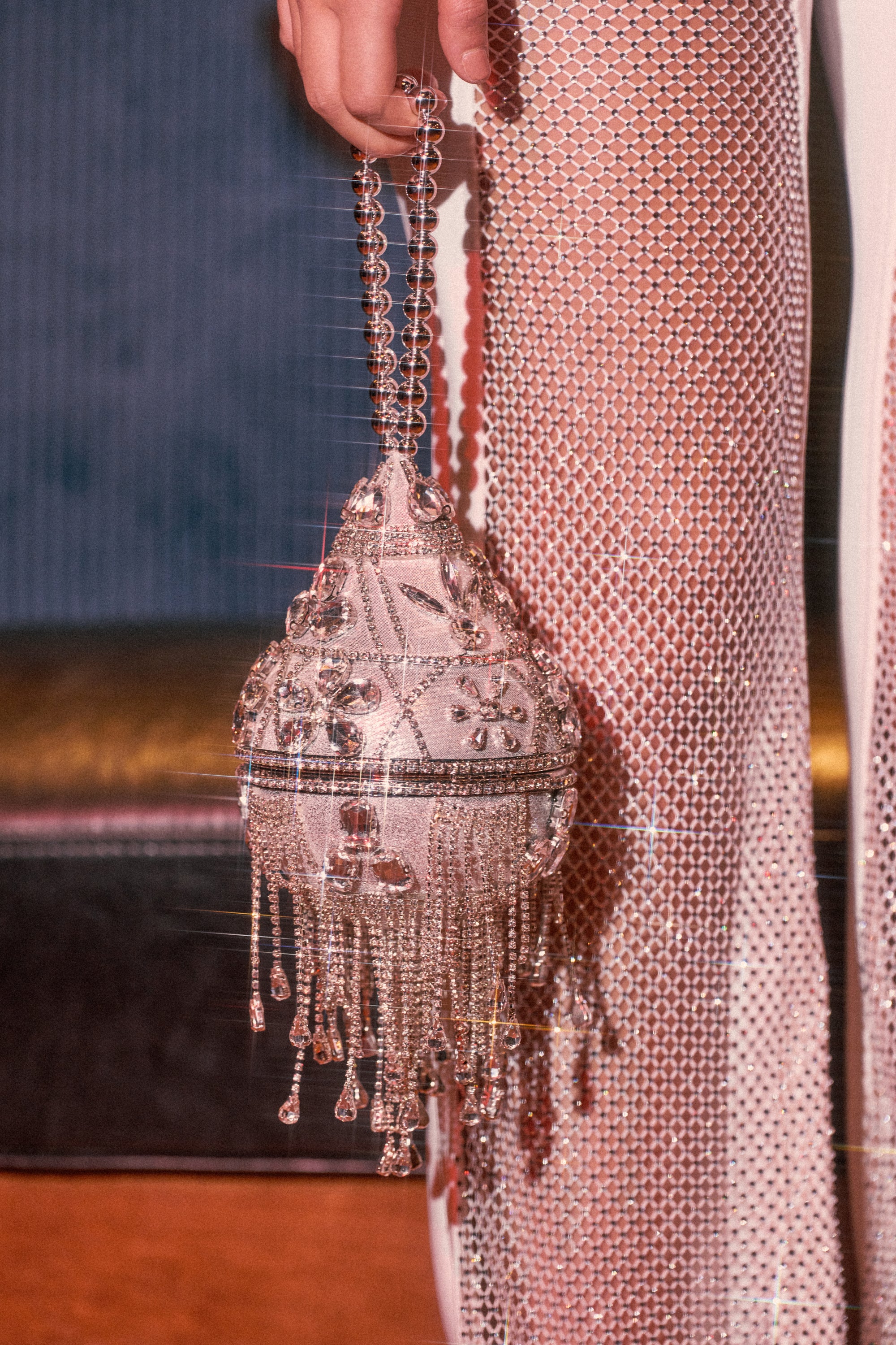 A hand holds the DECO BLING BAG, sparkling with ornate details and beaded fringe, while the person wears a shimmering rhinestone mesh dress—both catching the light beautifully.