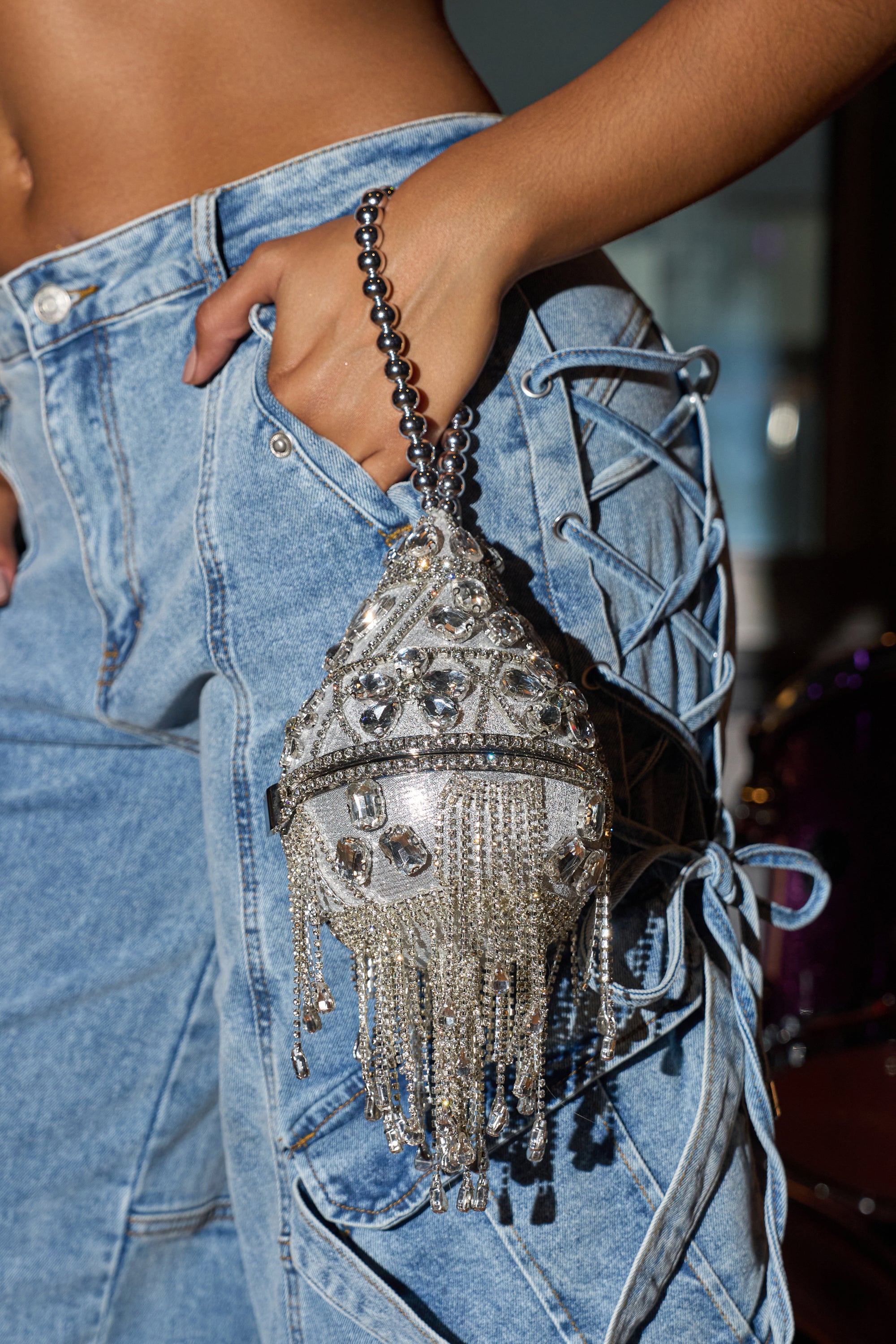 A person in lace-up, light-wash denim pants and a crop top holds the DECO BLING BAG—a dazzling crystal teardrop accessory embellished with rhinestones and dangling fringe accents. 0