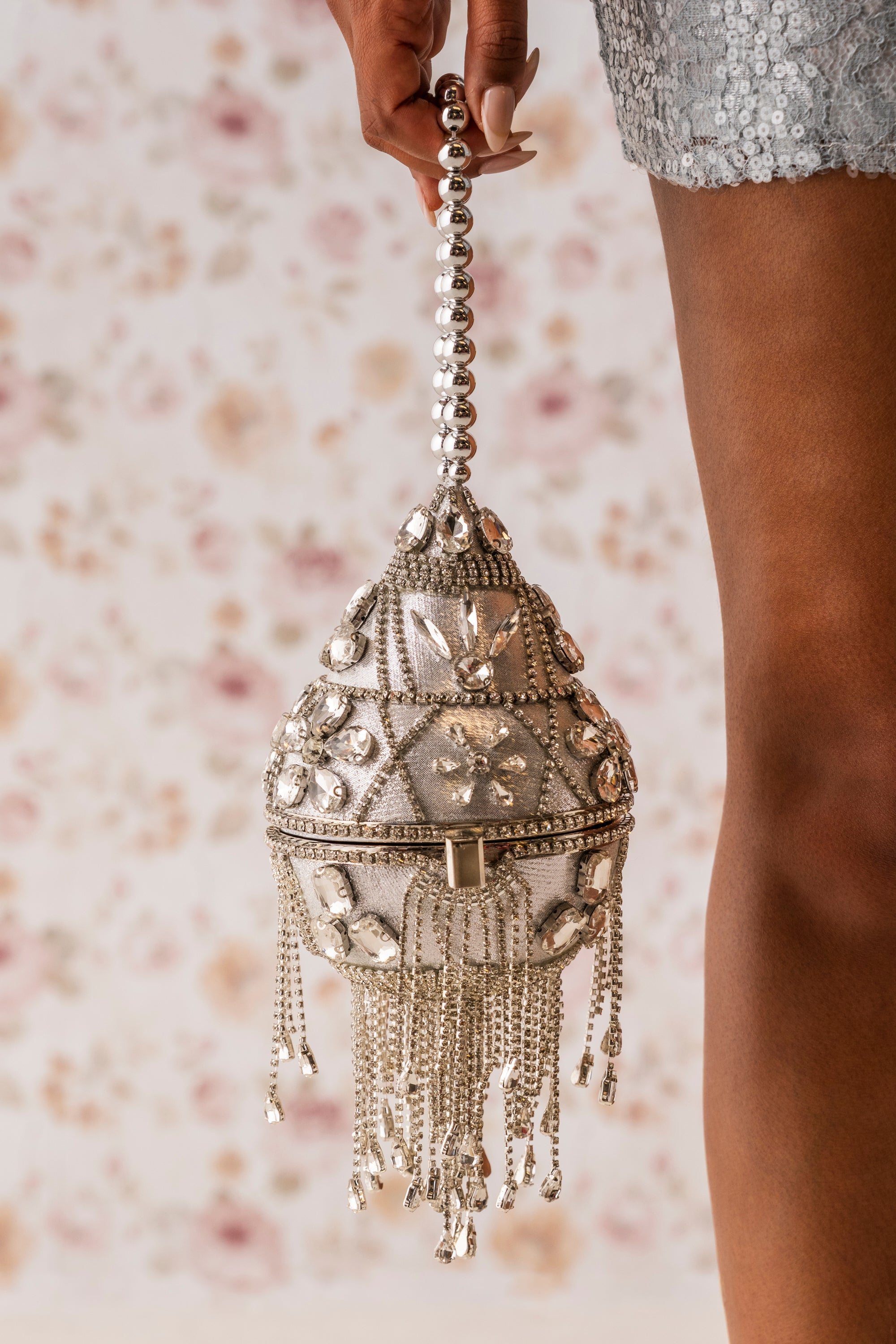 A person holds the DECO BLING BAG, a silver beaded rhinestone evening bag with chain fringe. The background is floral, and part of a sequined outfit plus the person's leg are visible. 1