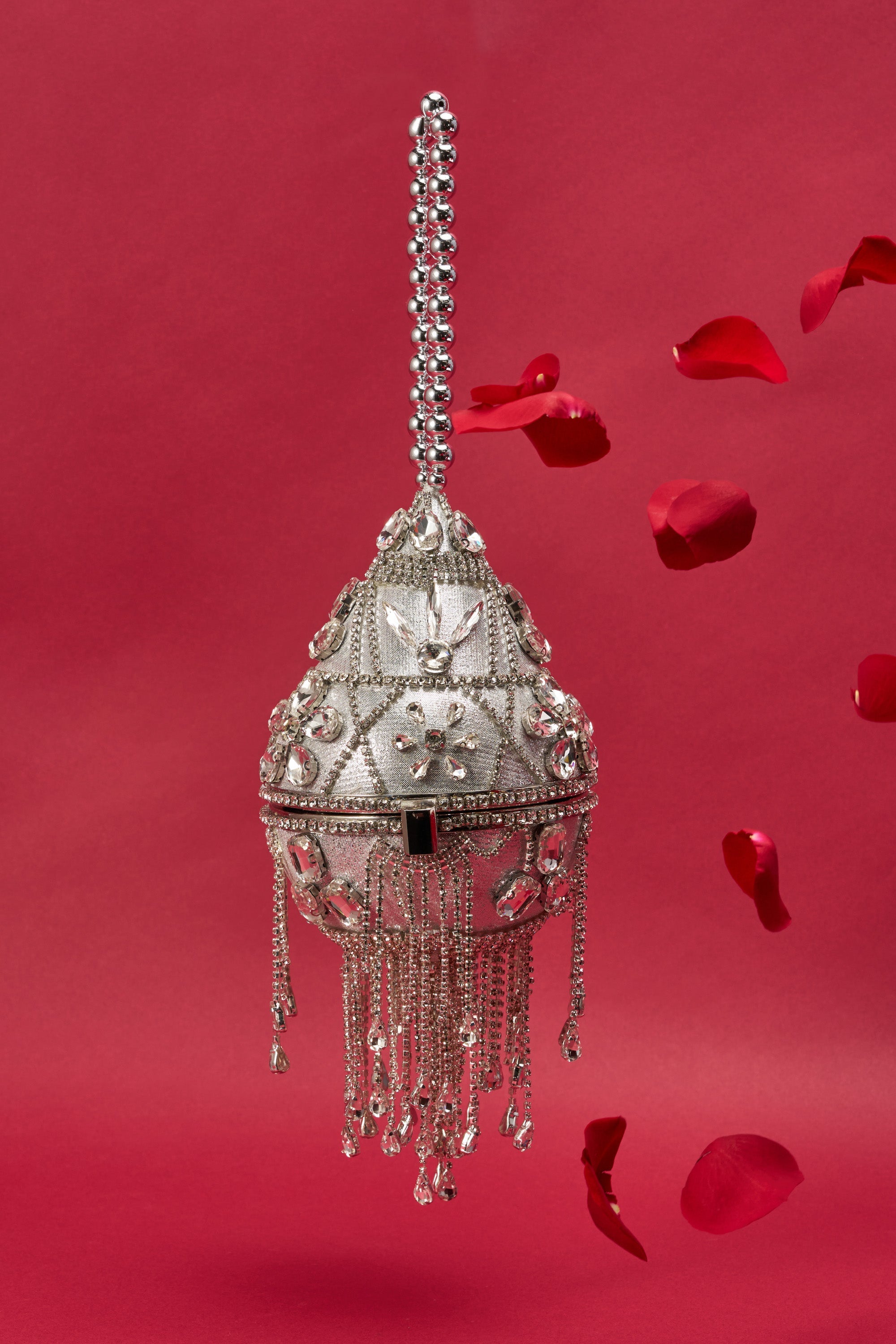The DECO BLING BAG, featuring crystal fringe, hanging chains, and intricate patterns, is displayed against a red background with floating red rose petals beside this striking accessory. 2