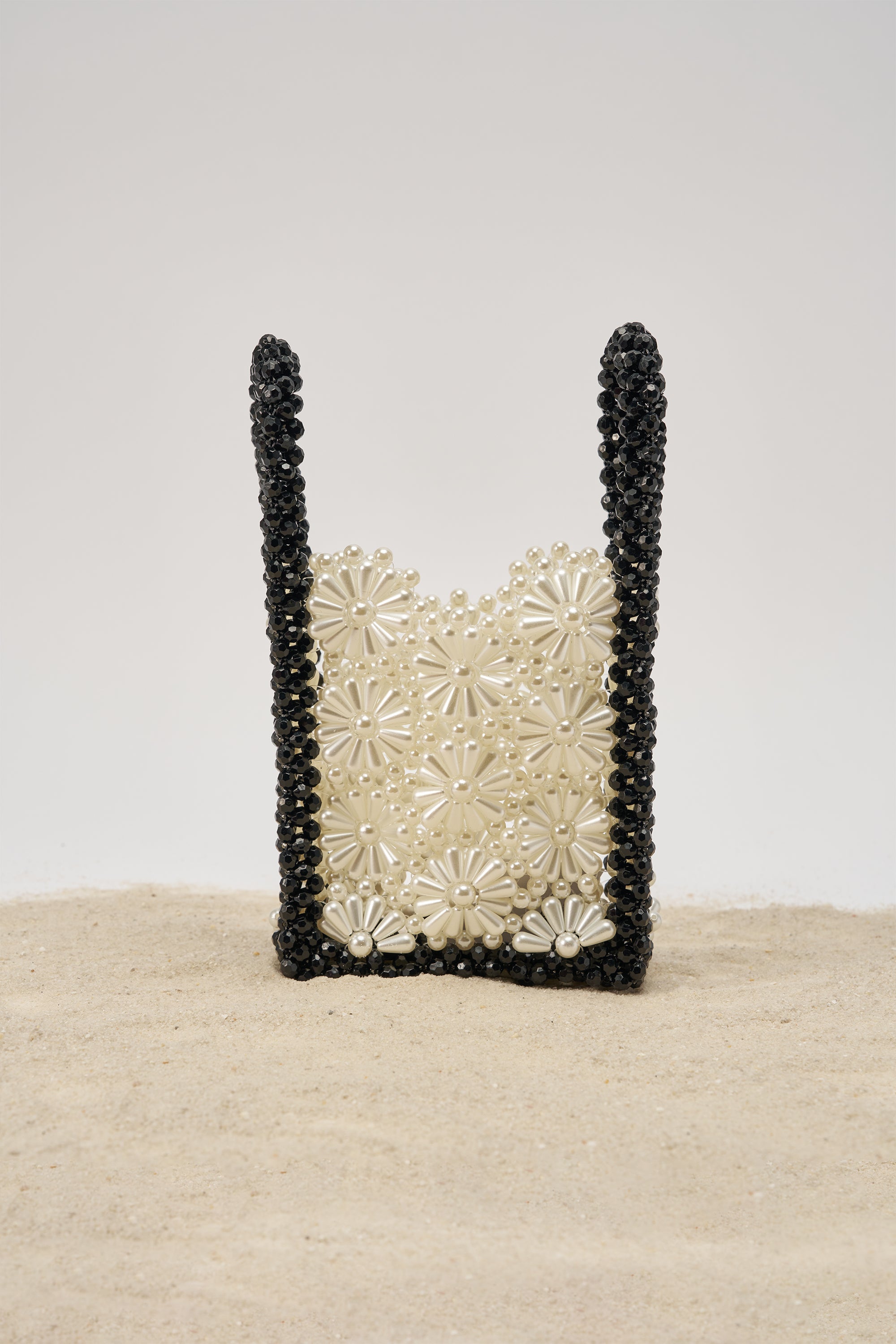 The ALL NIGHT LONG BEADED BAG, featuring black beaded handles and cream floral embellishments, stands upright on sand against a plain, light background. 0