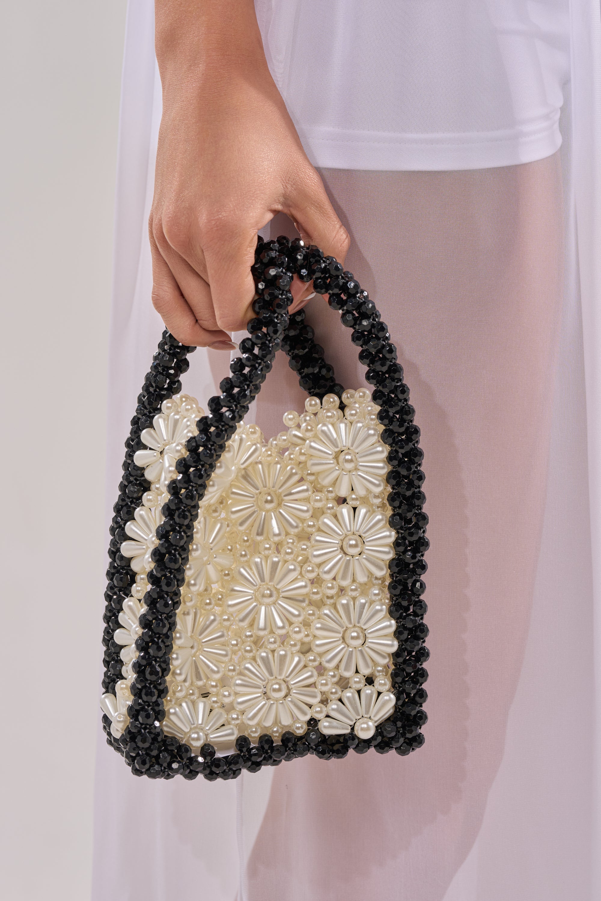 A hand holds the ALL NIGHT LONG BEADED BAG, featuring black handles and borders, pearl beading, and white floral patterns, set against a sheer white garment background. 2