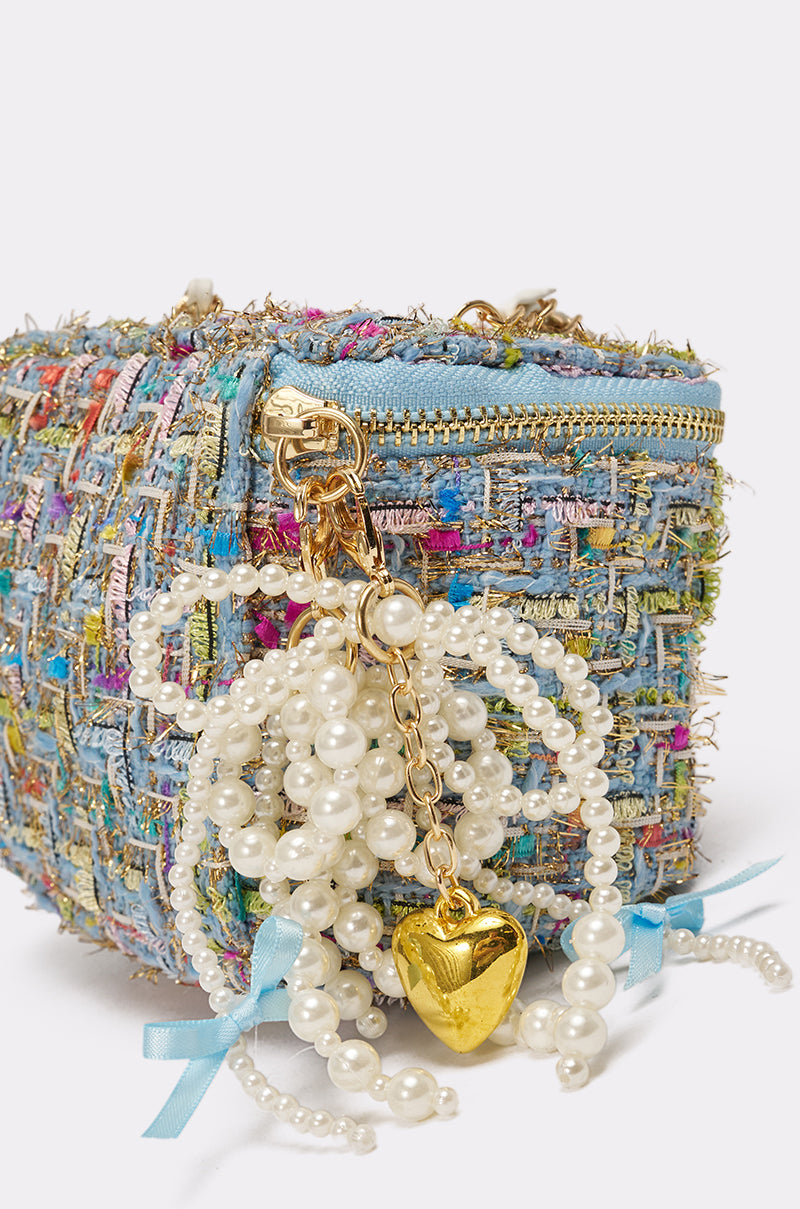 A close-up of the I HAVE CLASS TWEED PURSE, a multicolor woven handbag featuring a blue zipper, pearl beaded strands, gold heart charm, and blue ribbon bows as decorative accents. 5
