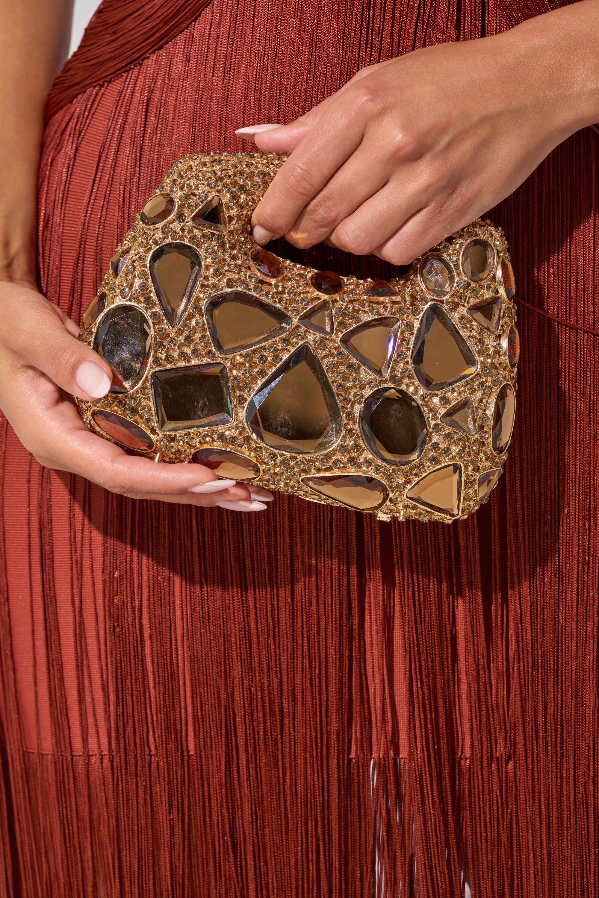 A person in a rust-colored fringe dress holds the EVA BLING BAG, a small gold evening bag adorned with mirrored and metallic studs. 0