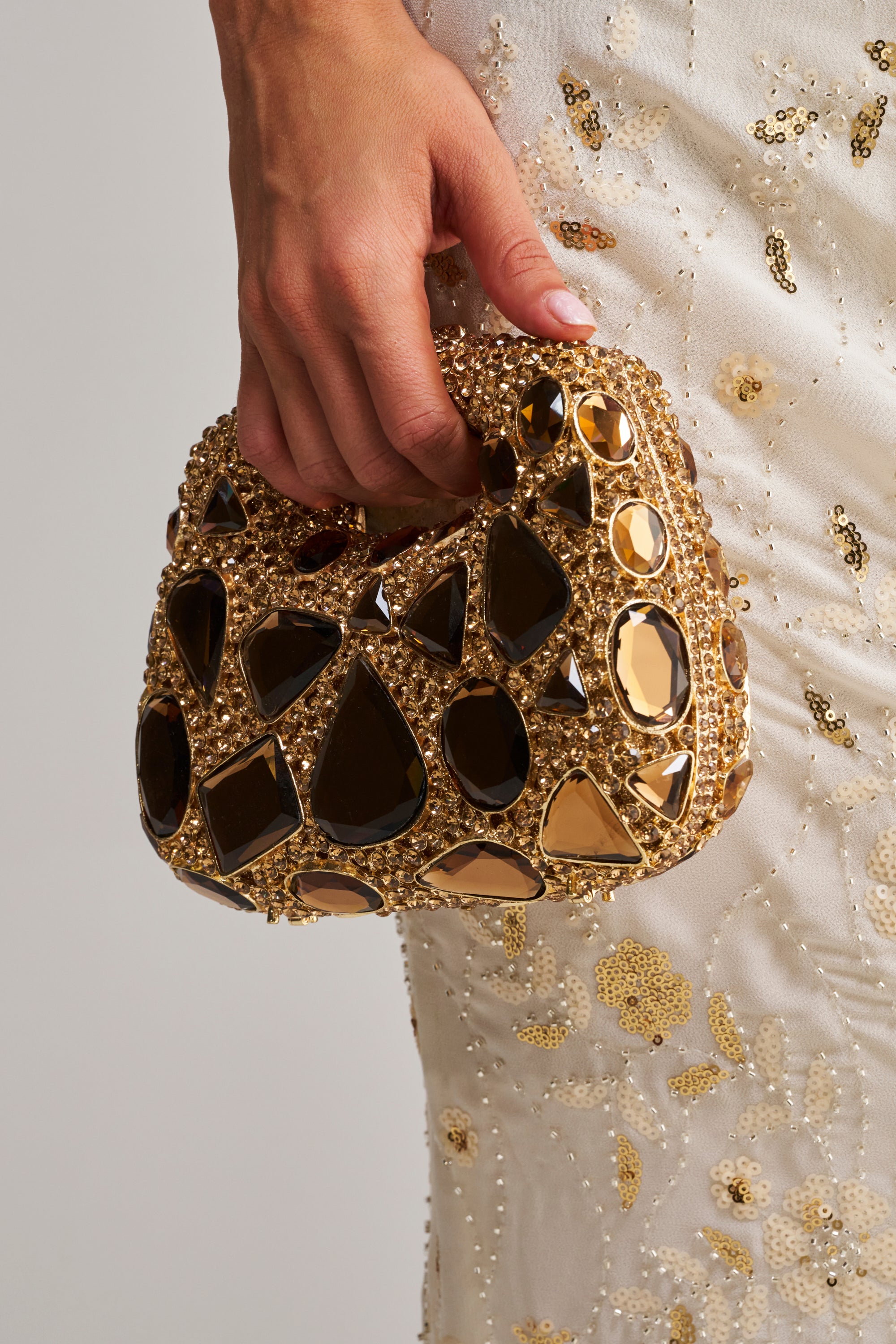 A hand holds the EVA BLING BAG, a mini evening bag embellished with large gold and black gemstones, perfectly complementing a white beaded dress with intricate floral patterns.
