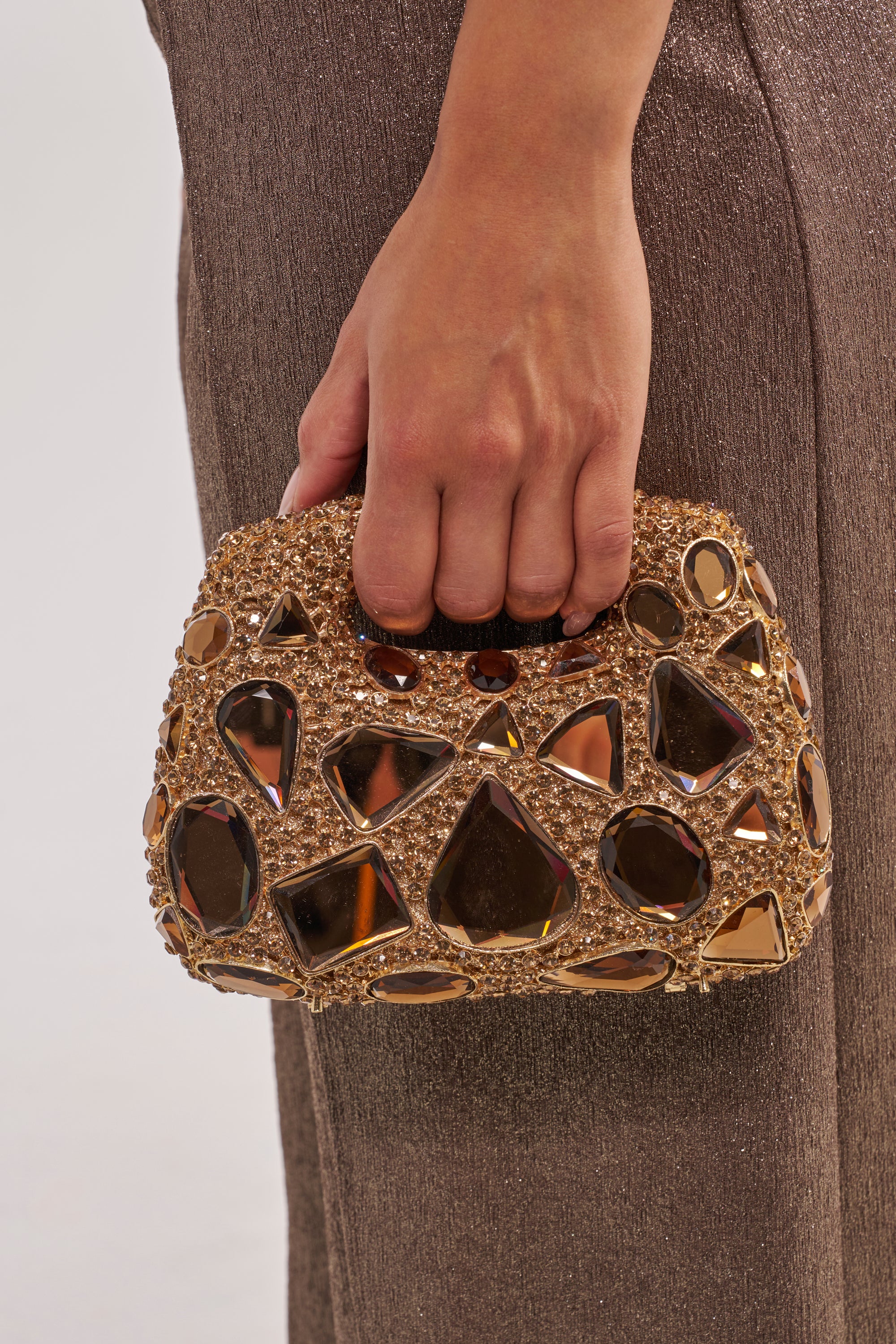 A person in shimmery taupe pants holds the EVA BLING BAG, a rhinestone mini handbag featuring mirrored geometric shapes, gemstone accents, and an elegant gold chain strap. 3