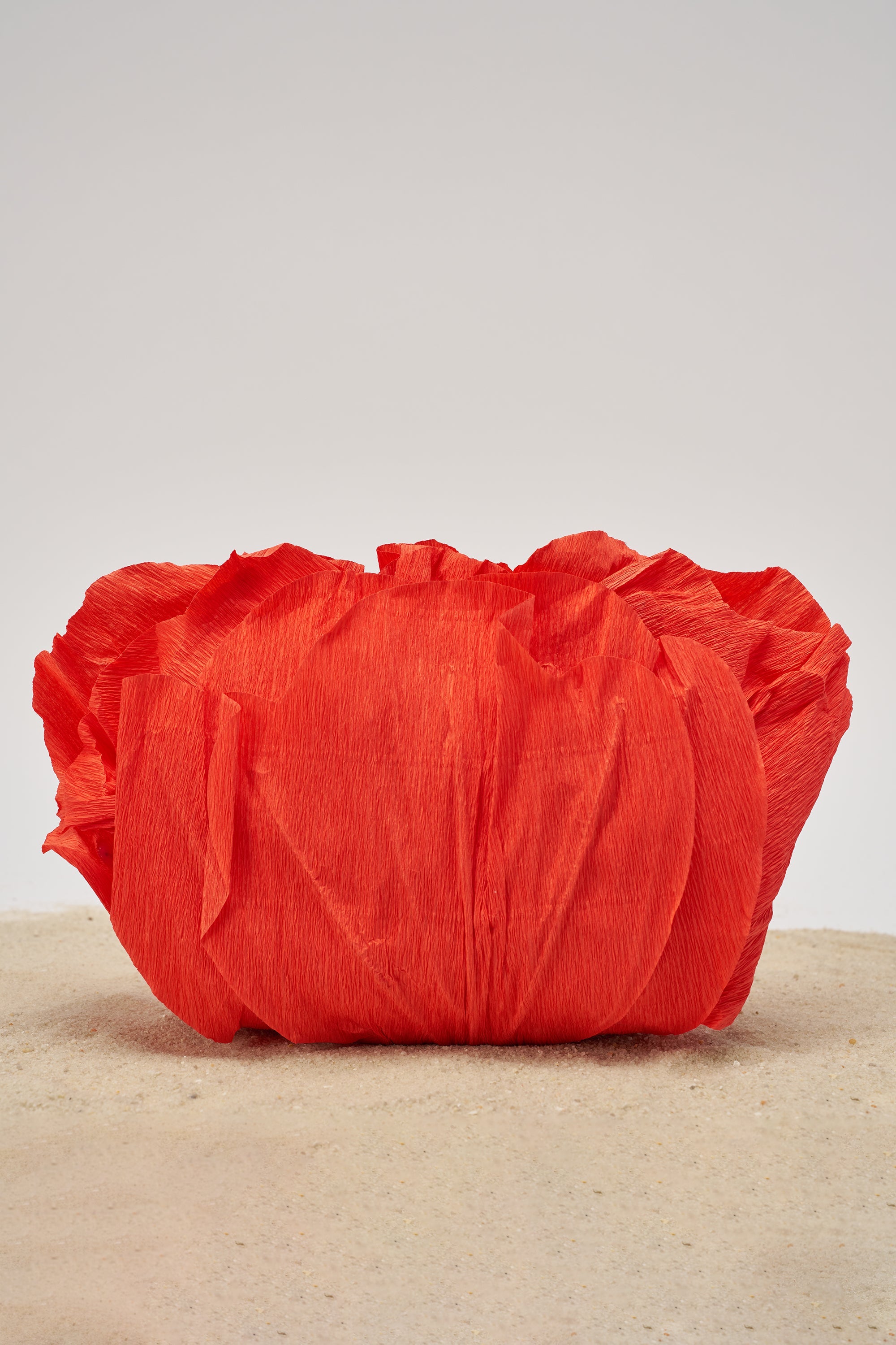 The FEEL MY VIBE CLUTCH, a bright red, textured evening bag with a sculptural design, rests on a sandy surface against a plain light background. 1