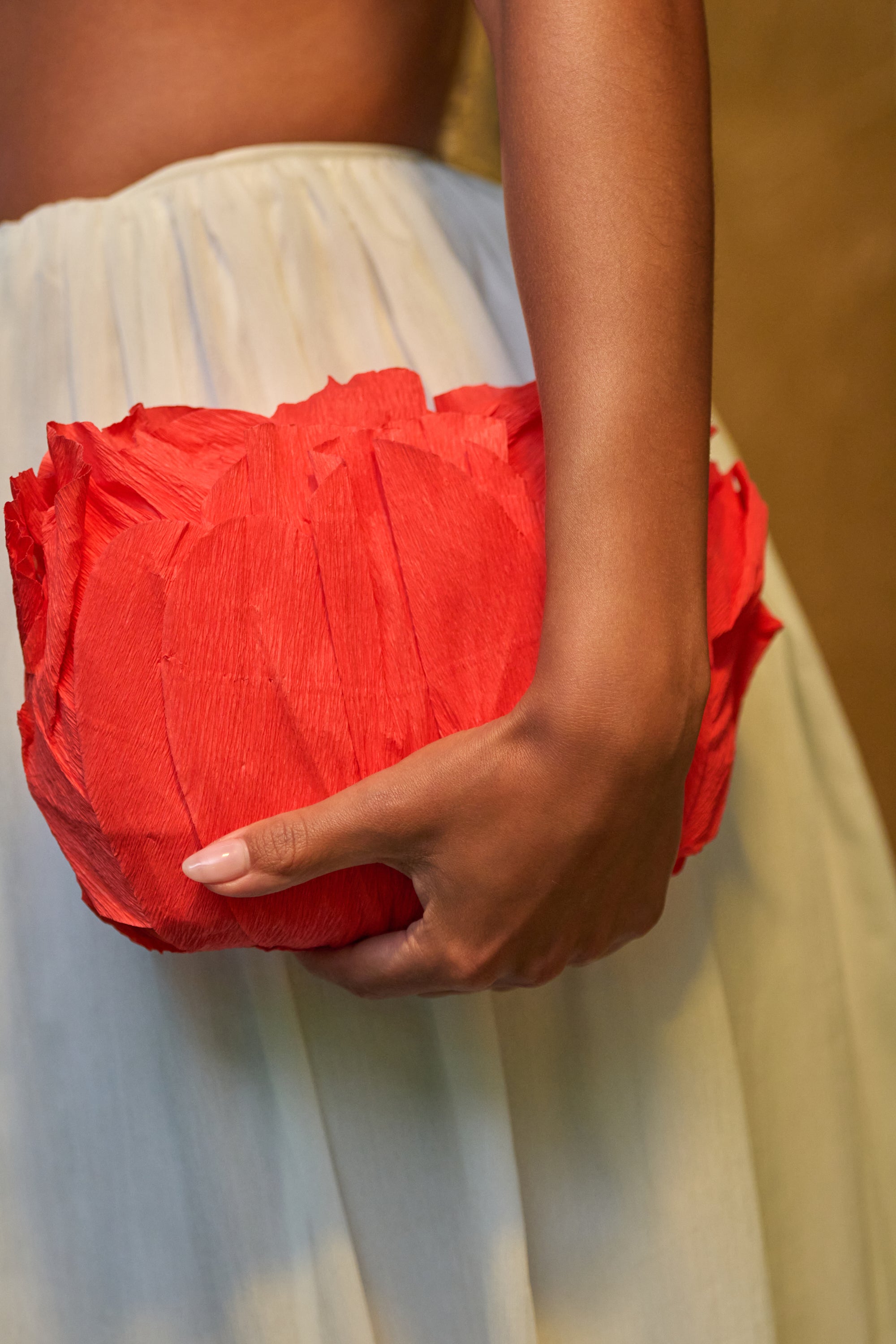 A person in a white skirt holds the FEEL MY VIBE CLUTCH, a vibrant orange bag with a petal-like texture that stands out as a true statement piece against the softly blurred background. 0