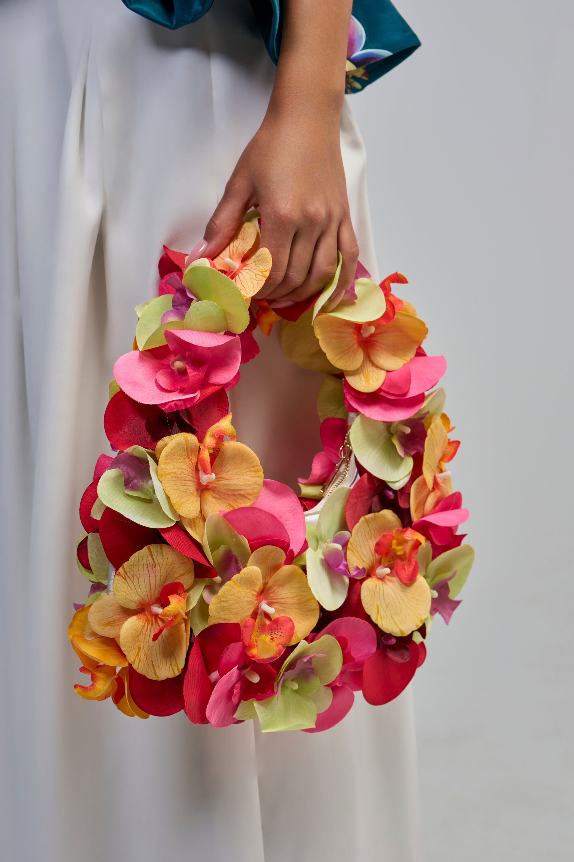 A hand holds the ORCHID LOVE PURSE, a vibrant 3D floral handbag adorned with colorful artificial flowers in shades of pink, yellow, orange, and green, shown against a light background. The person is wearing white pants.