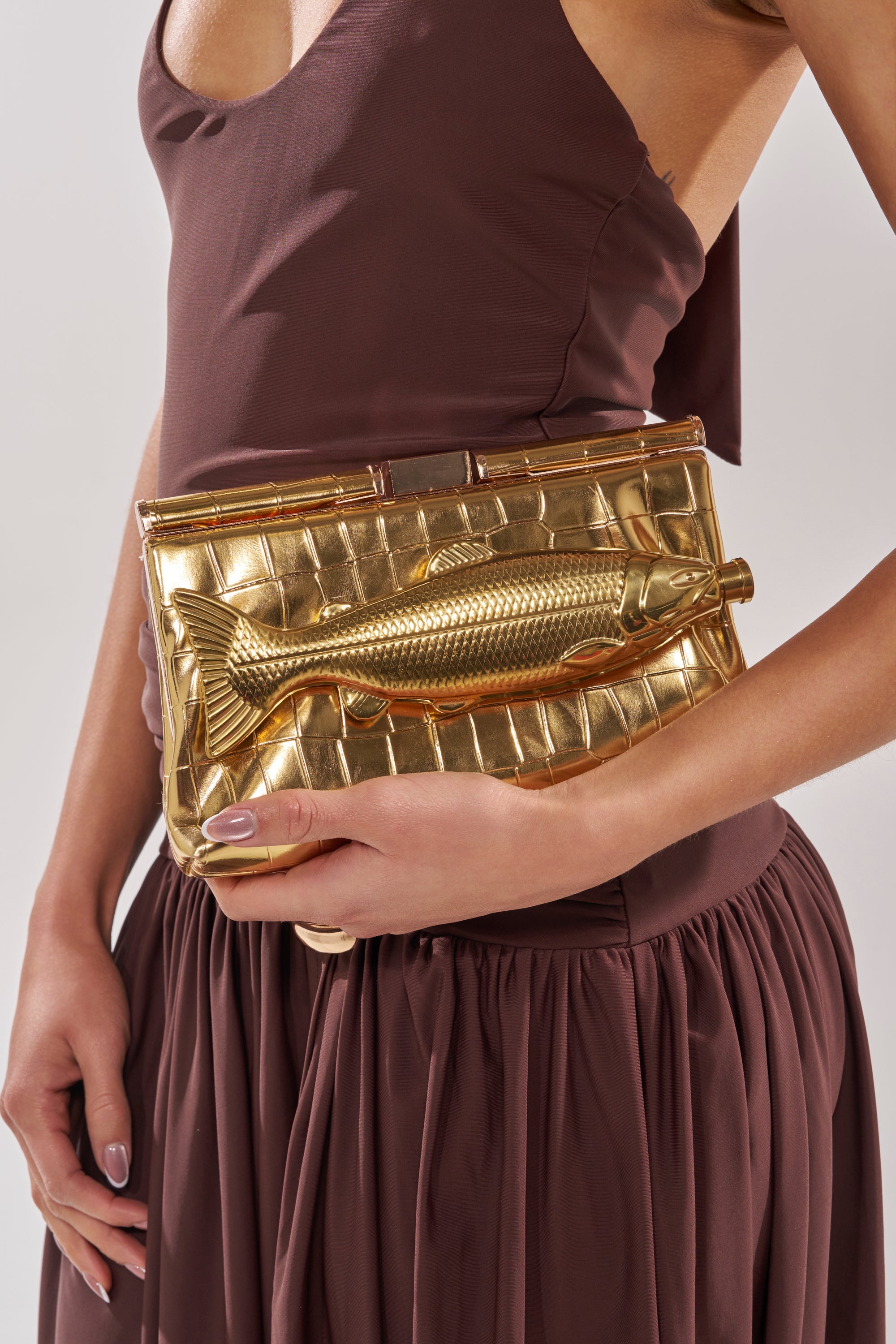 A person in a sleeveless brown dress holds the GONE FISHIN CLUTCH, a metallic faux leather accessory featuring a raised fish design. 3