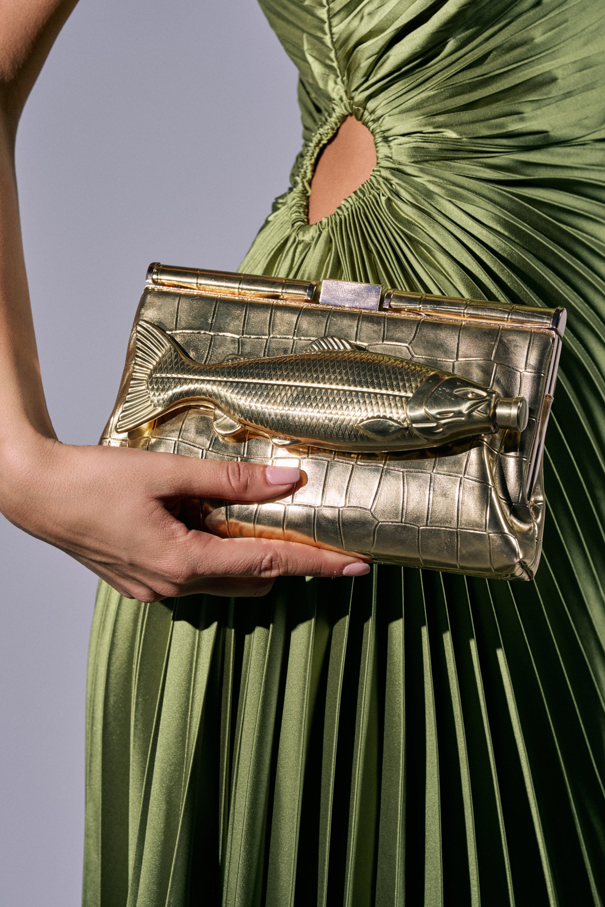 A person in a pleated light green dress holds the GONE FISHIN CLUTCH, a gold metallic faux leather bag with a raised fish design and removable crossbody chain. The background is plain and softly lit. 1
