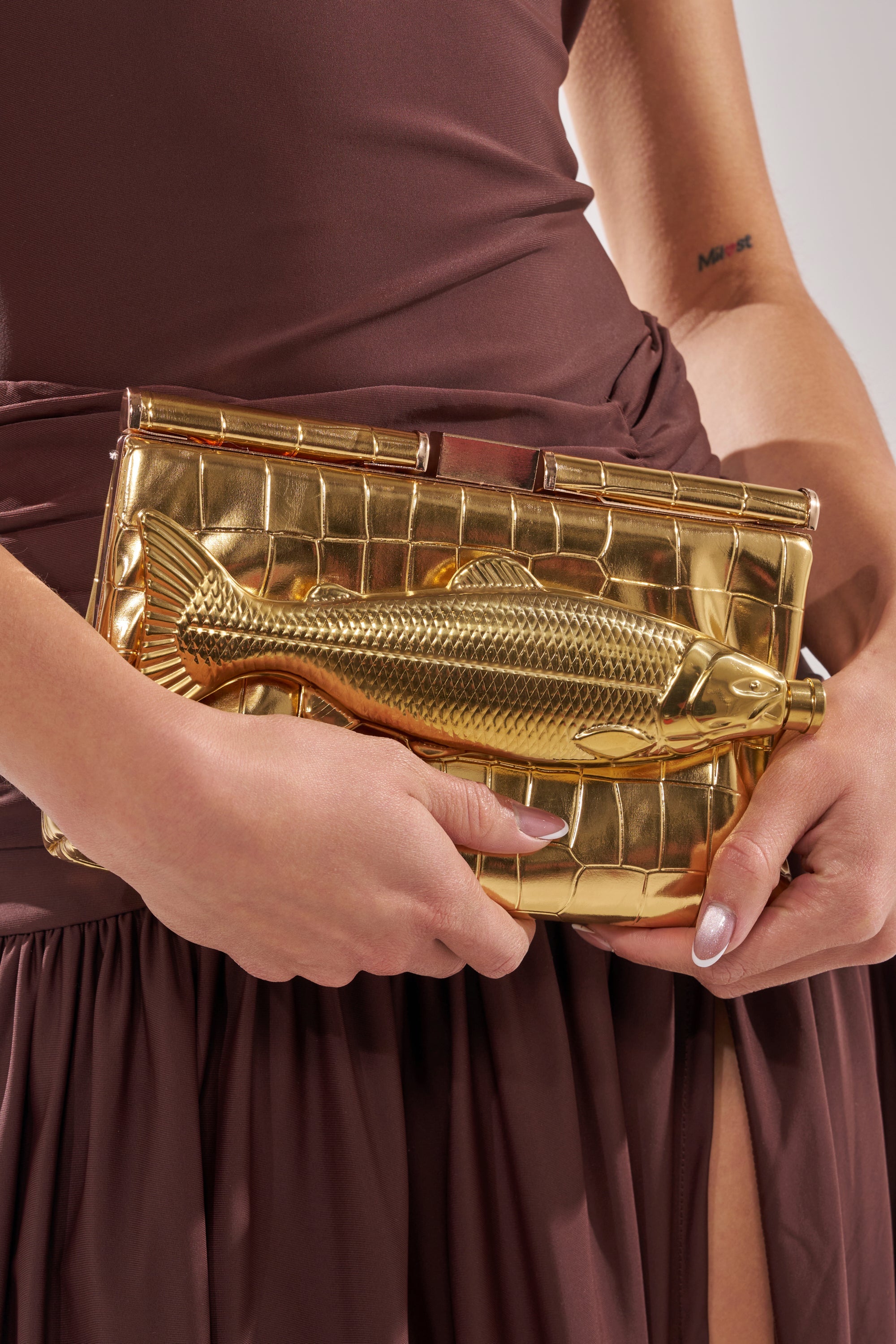 A person in a mauve dress holds the GONE FISHIN CLUTCH, a gold croc-embossed metallic faux leather bag with a sculpted fish detail and removable crossbody chain; a small inner arm tattoo is visible. 7