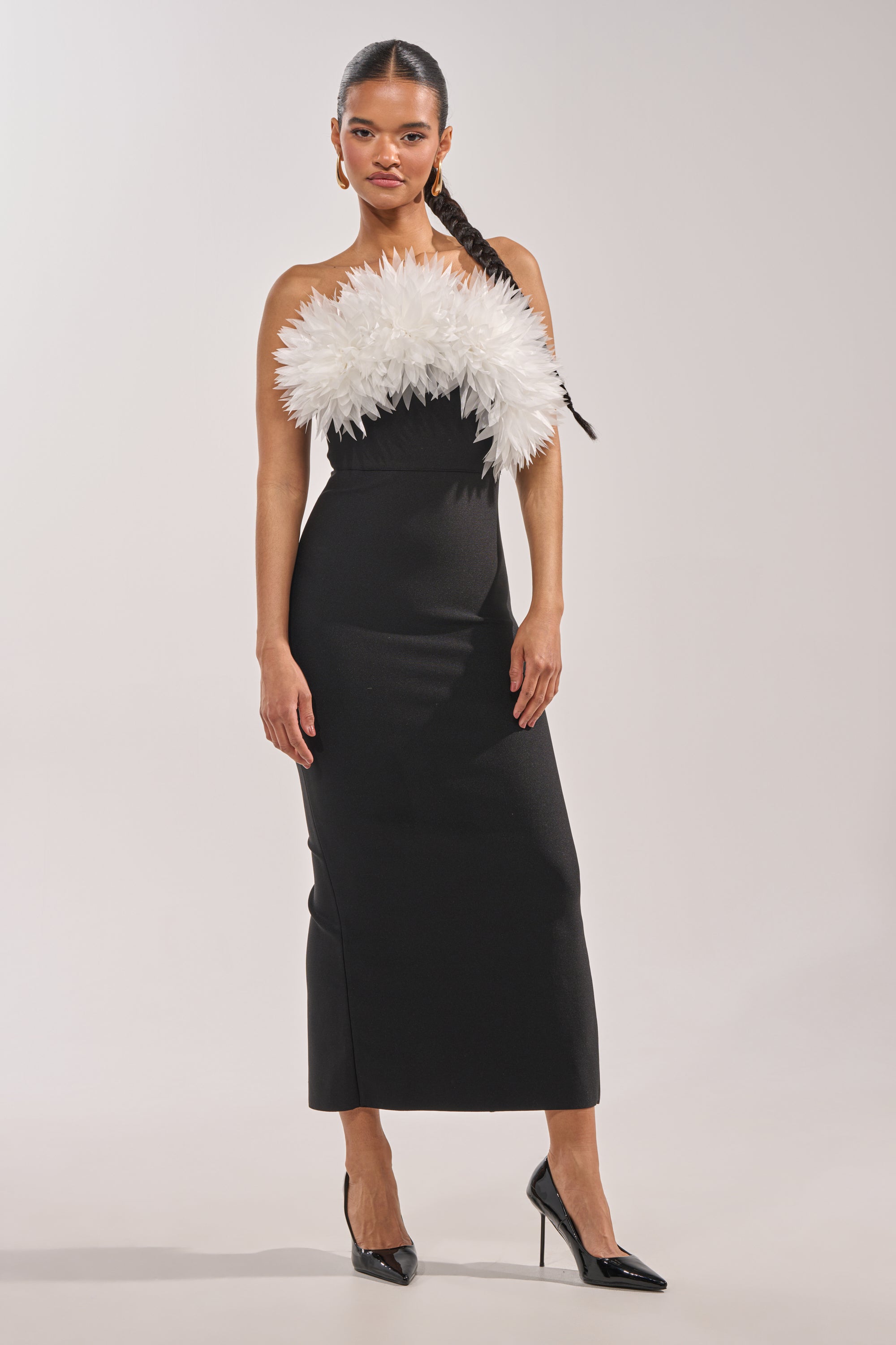 A woman poses in the HEART ON MY SLEEVE BANDAGE MIDI DRESS IN BLACK, featuring a strapless design with striking white feathered detail across the chest. She pairs it with black heels and a long braid, set against a plain light background. 1